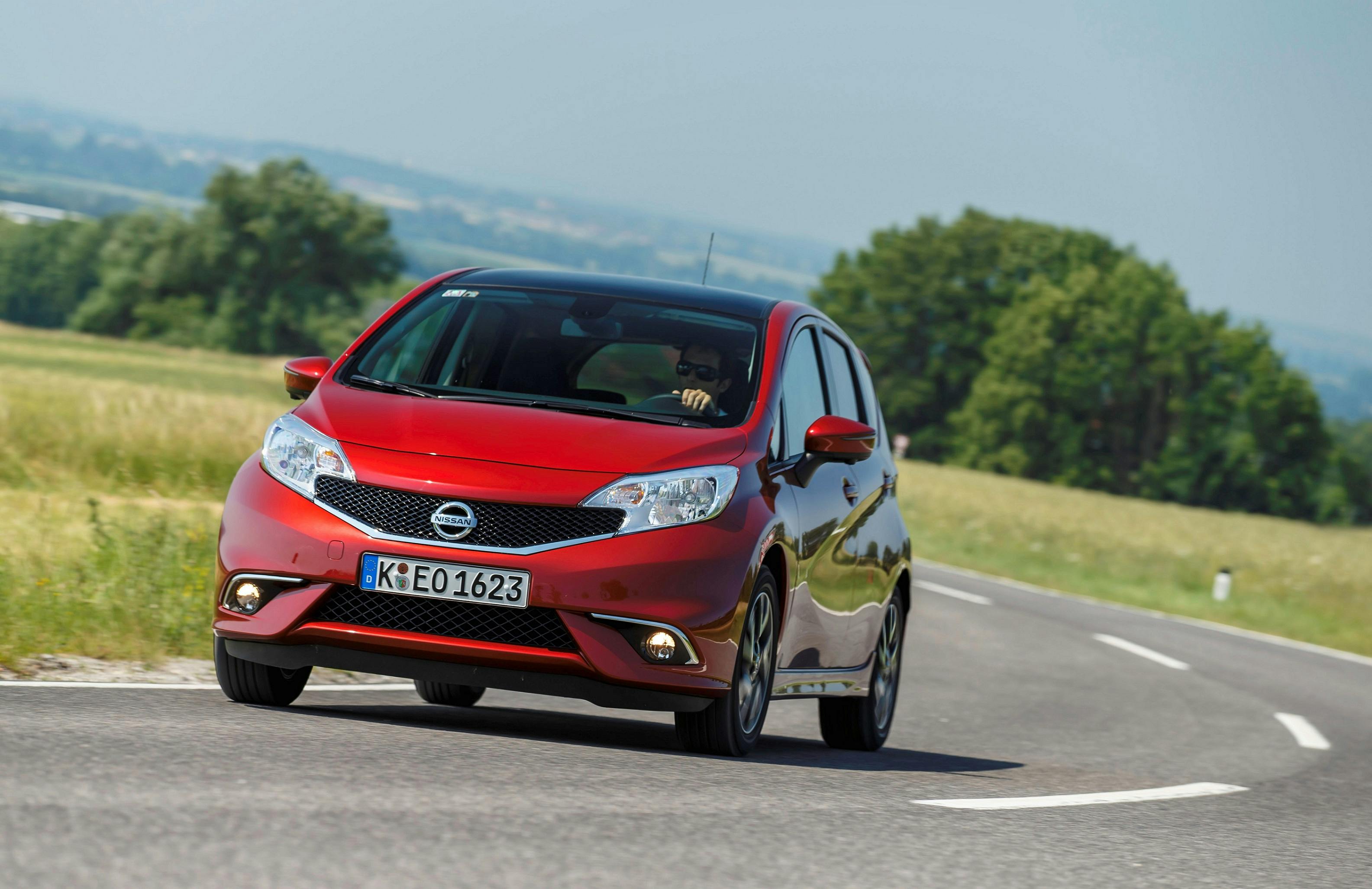 Roter Nissan Note auf einer Landstraße, umgeben von grünen Wiesen und Bäumen, bei sonnigem Wetter.