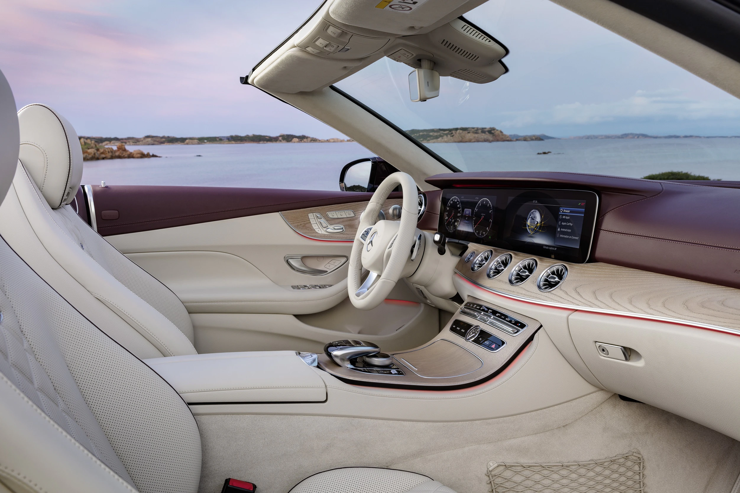 Luxuriöser Autoinnenraum mit cremefarbenen und weinroten Sitzen. Blick auf ein modernes Armaturenbrett und Touchscreen-Anzeigen. Meer im Hintergrund.