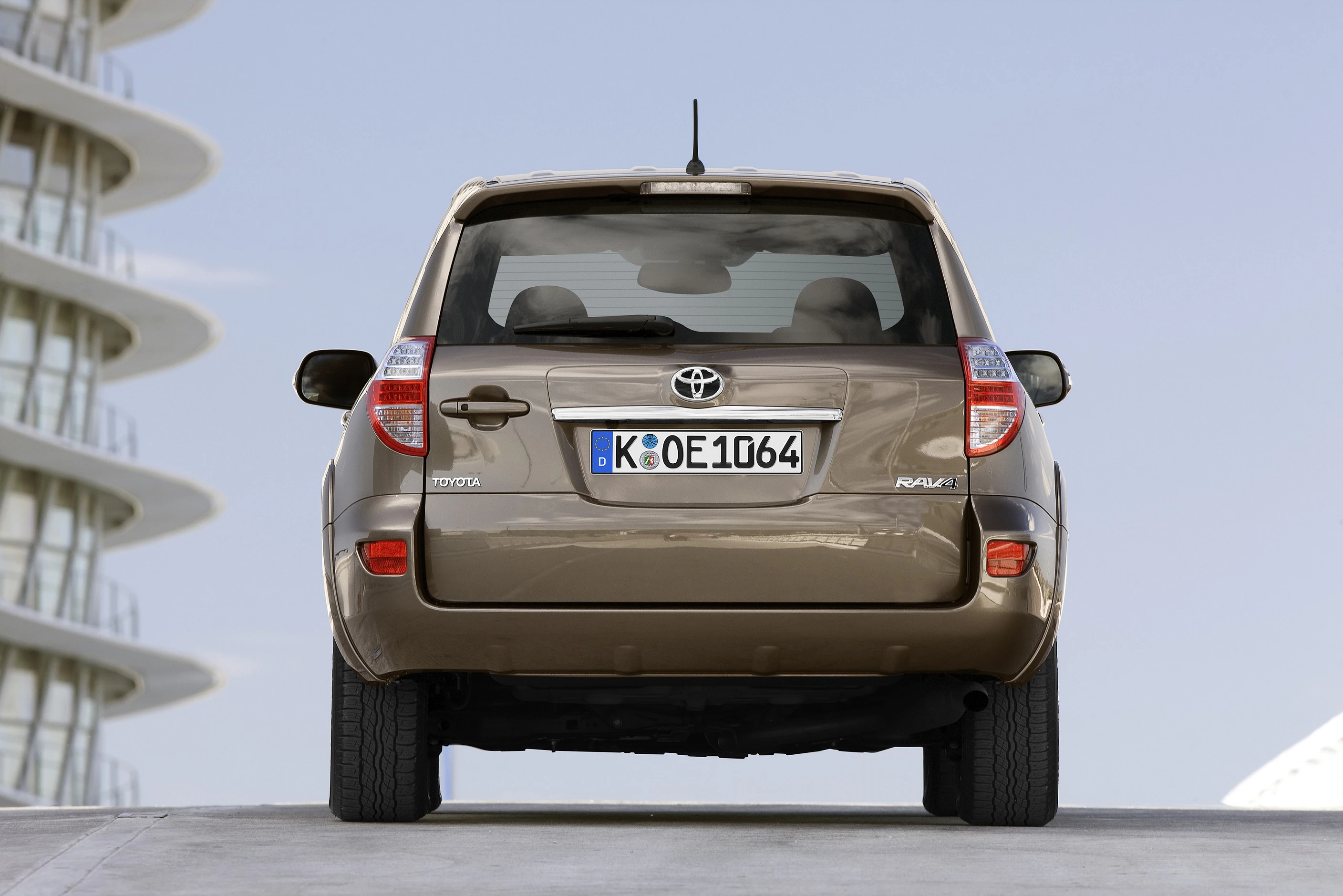 Ein brauner Toyota RAV4 von hinten, geparkt auf einem Betonweg, mit einem modernen Gebäude im Hintergrund und blauem Himmel.