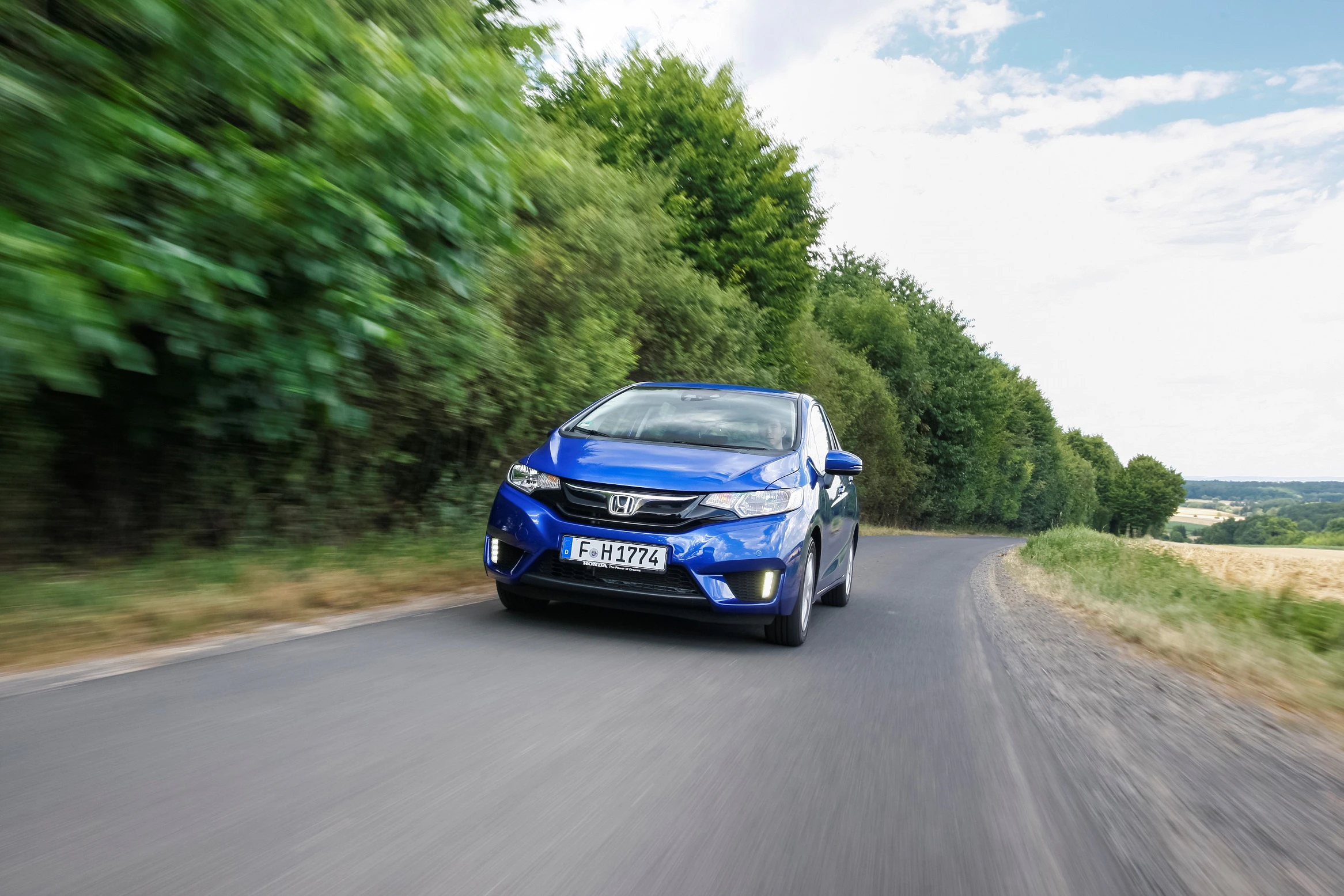 Blauer Honda fährt auf einer schmalen Landstraße, umgeben von grünen Bäumen und Wiesen unter blauem Himmel.