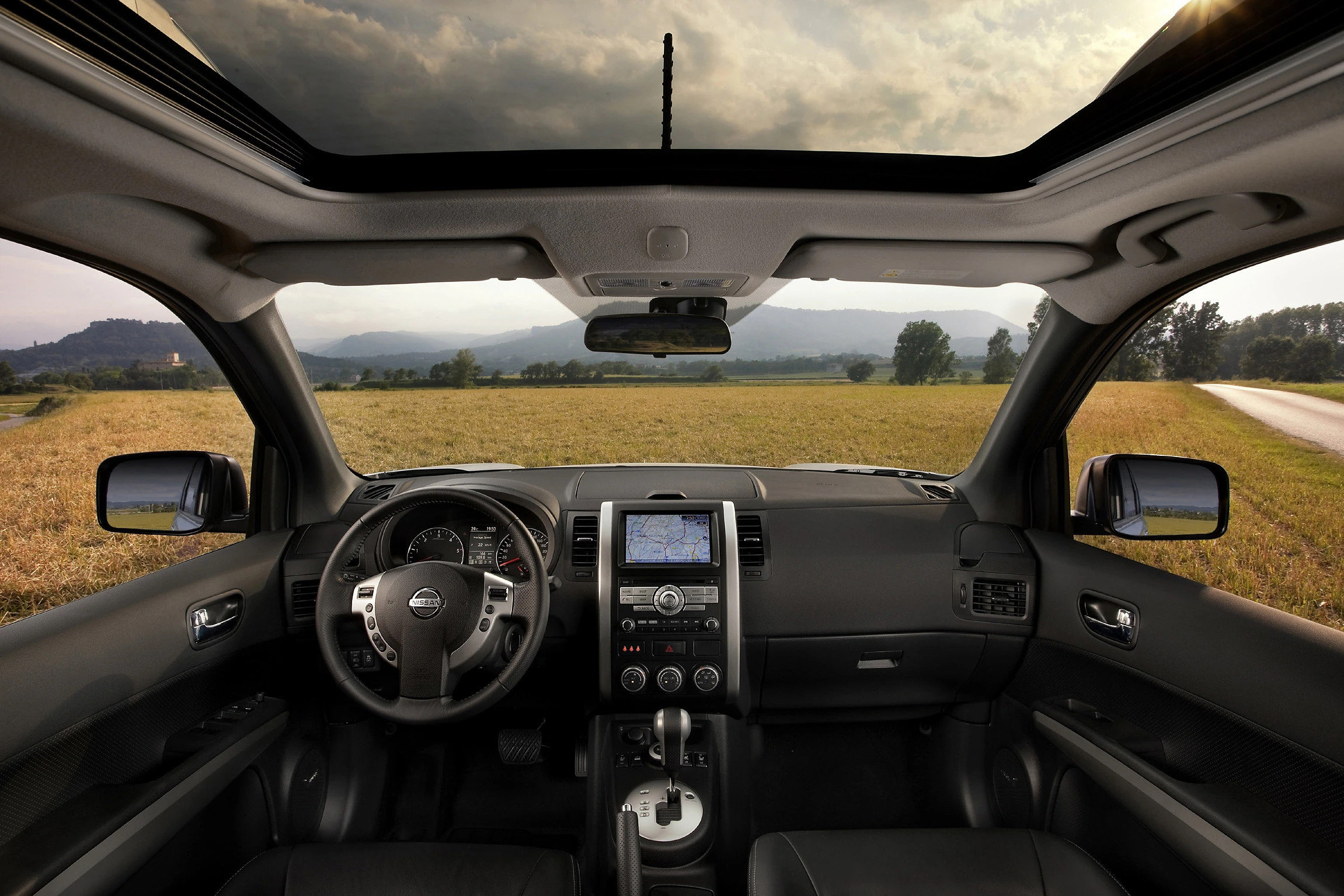 Innenansicht eines Autos mit Panoramadach, Blick auf Lenkrad, Armaturenbrett, und Navigation, draußen Felder und bewölkter Himmel.