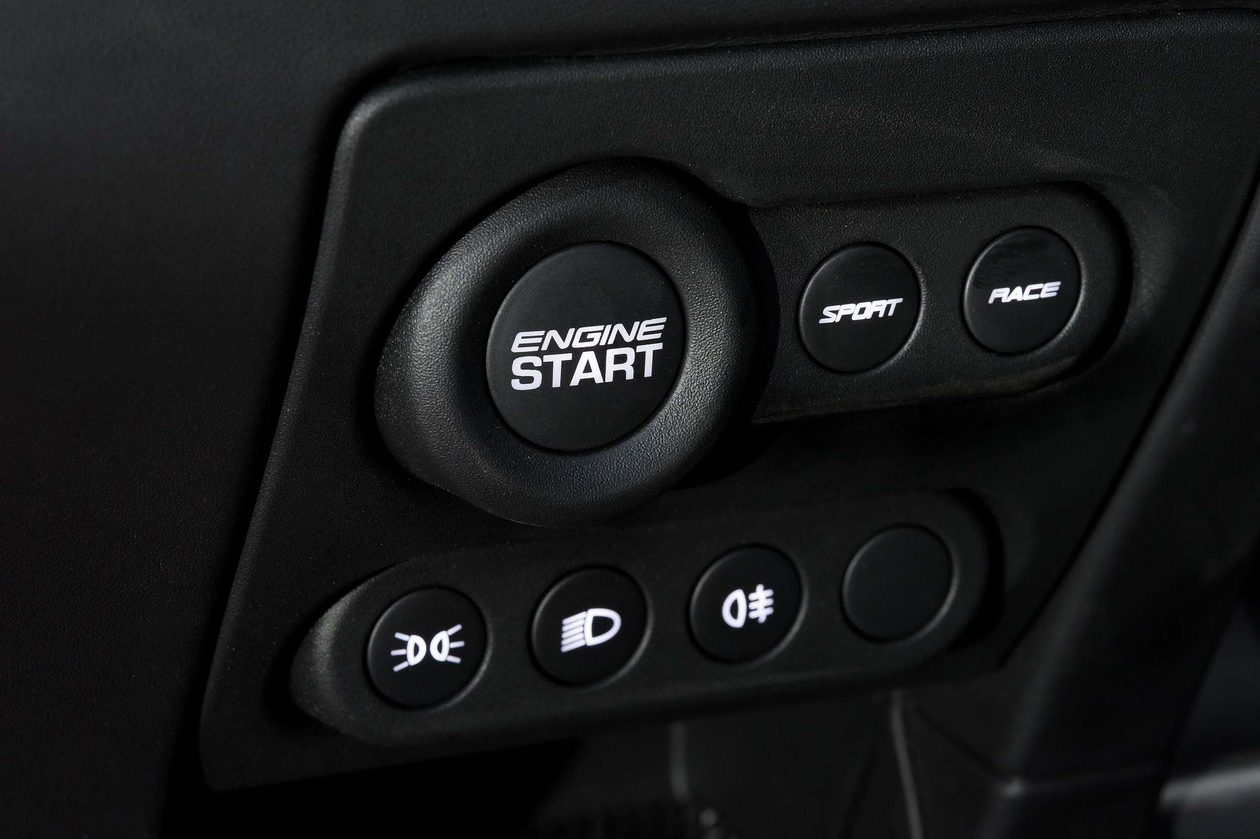 Nahaufnahme eines Auto-Startknopfs mit den Zusatzknöpfen „Sport“, „Race“ und Lichtsteuerfunktionen auf einem schwarzen Armaturenbrett.