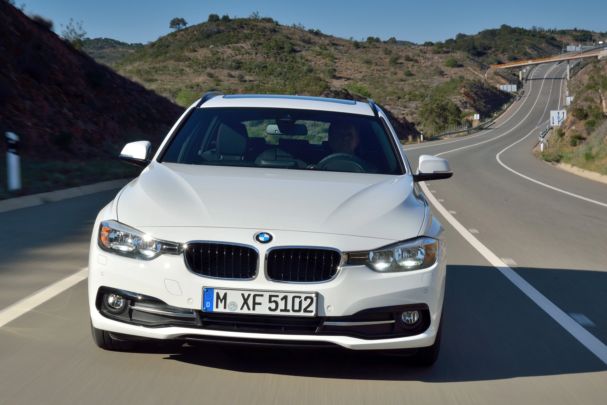 Ein weißer BMW fährt auf einer kurvenreichen Landstraße durch eine bergige Landschaft bei klarem Himmel.