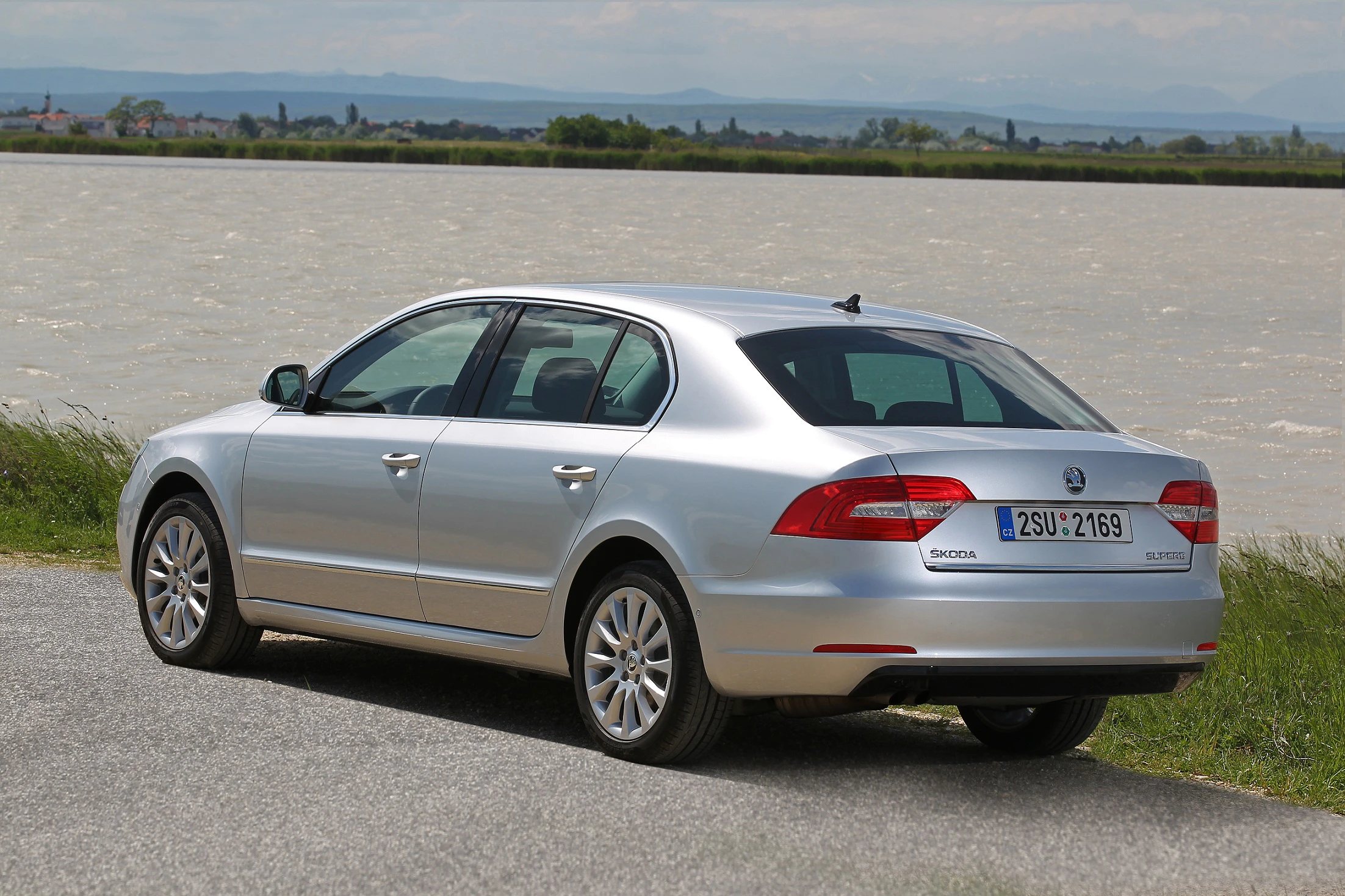 Silberner Skoda Superb parkt am Ufer eines Flusses mit grasbewachsenem Rand. Im Hintergrund sind Hügel sichtbar.