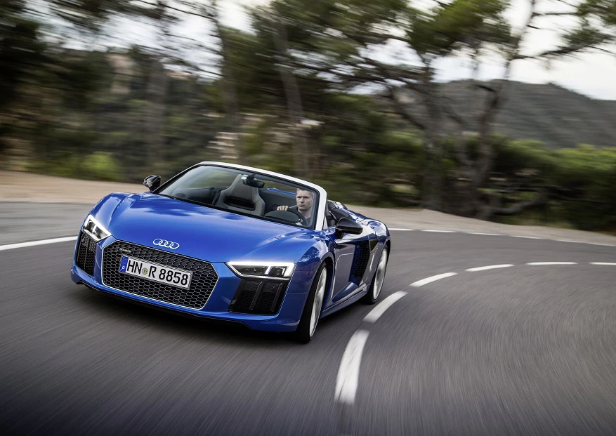 Blauer Audi-Sportwagen in Kurvenfahrt auf Landstraße, bei Tag und schneller Fahrt. Bäume und verschwommene Landschaft im Hintergrund.