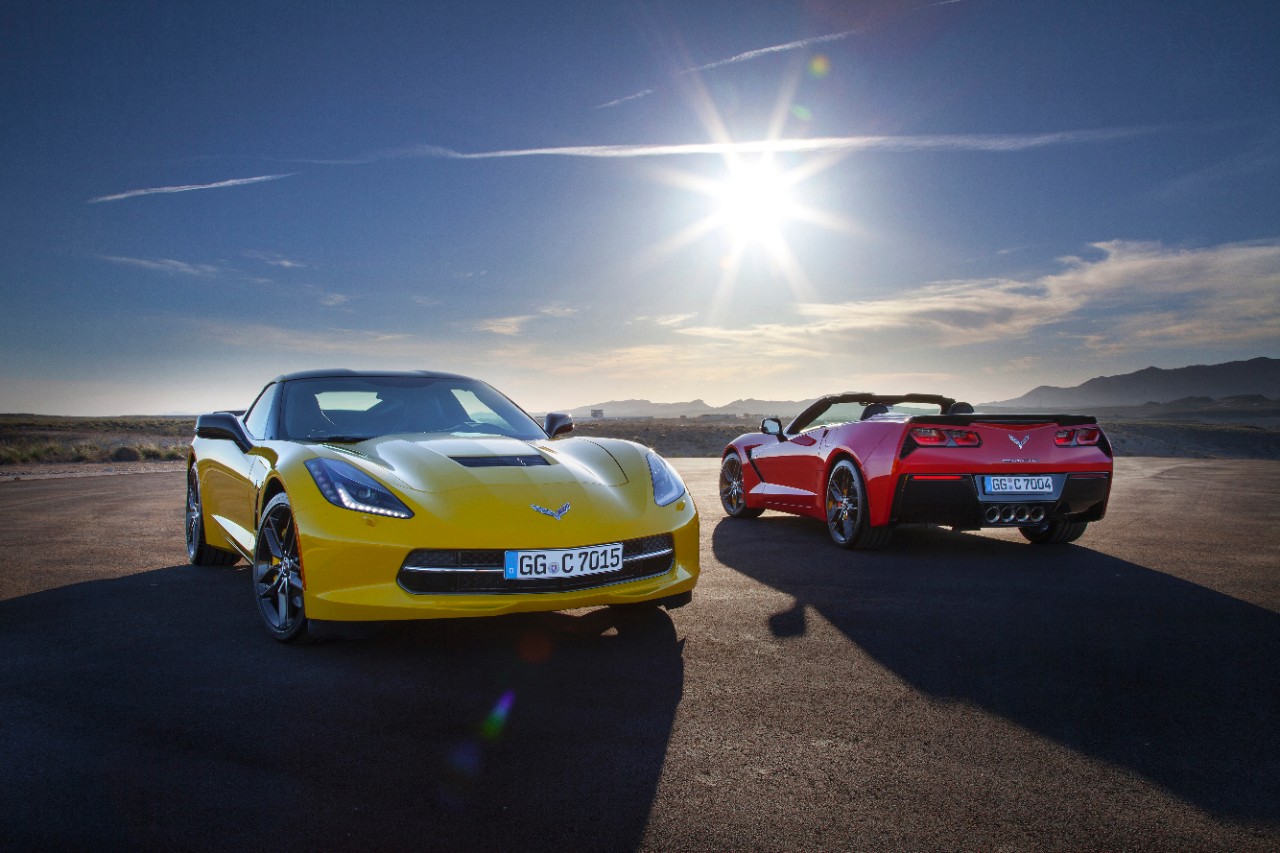 Zwei Sportwagen auf offener Straße bei Sonnenuntergang, ein gelbes Coupé und ein rotes Cabrio. Weitläufige Landschaft im Hintergrund.