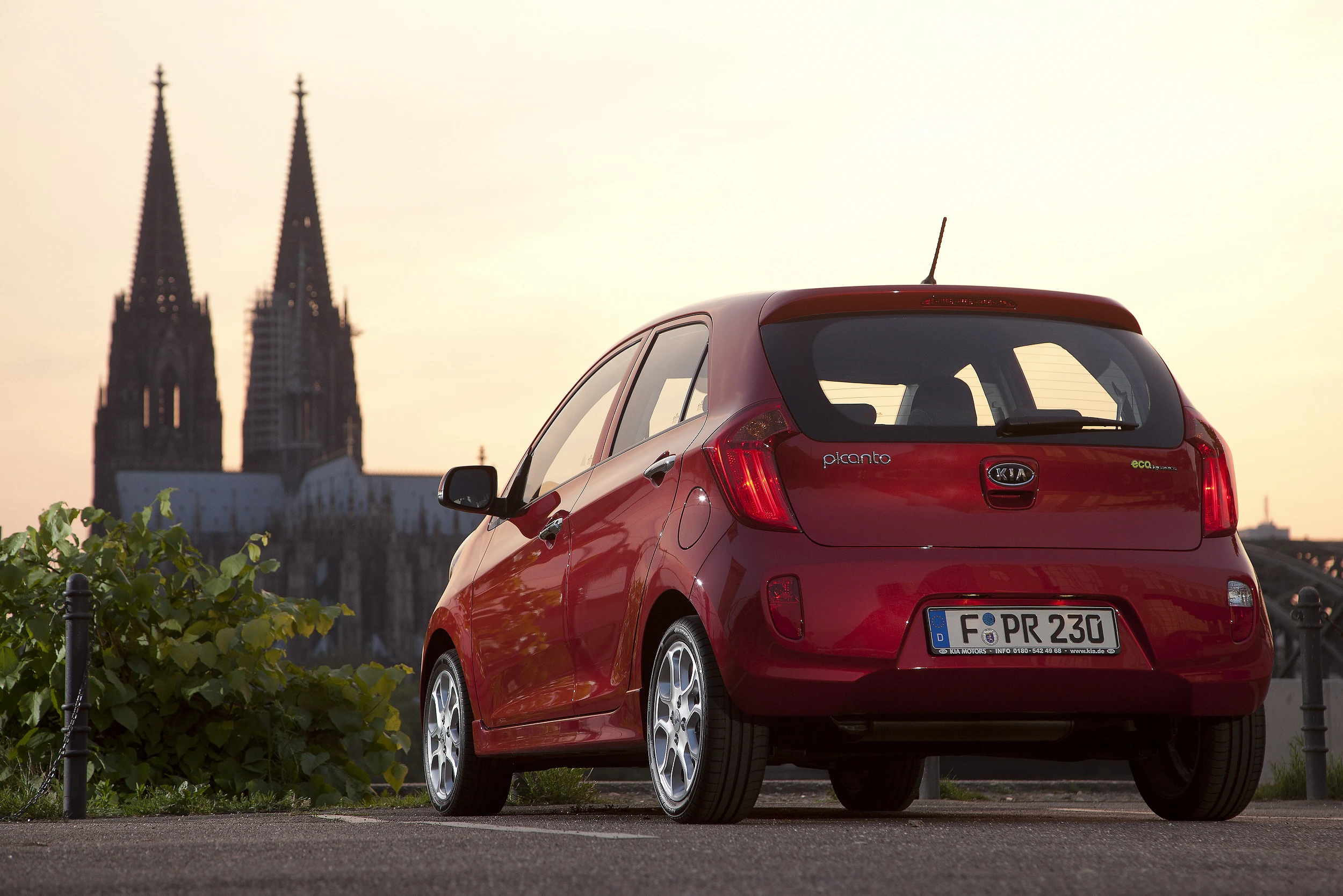 Roter Kia Picanto vor Kölner Dom bei Sonnenuntergang, perspektivisch von hinten rechts aufgenommen.