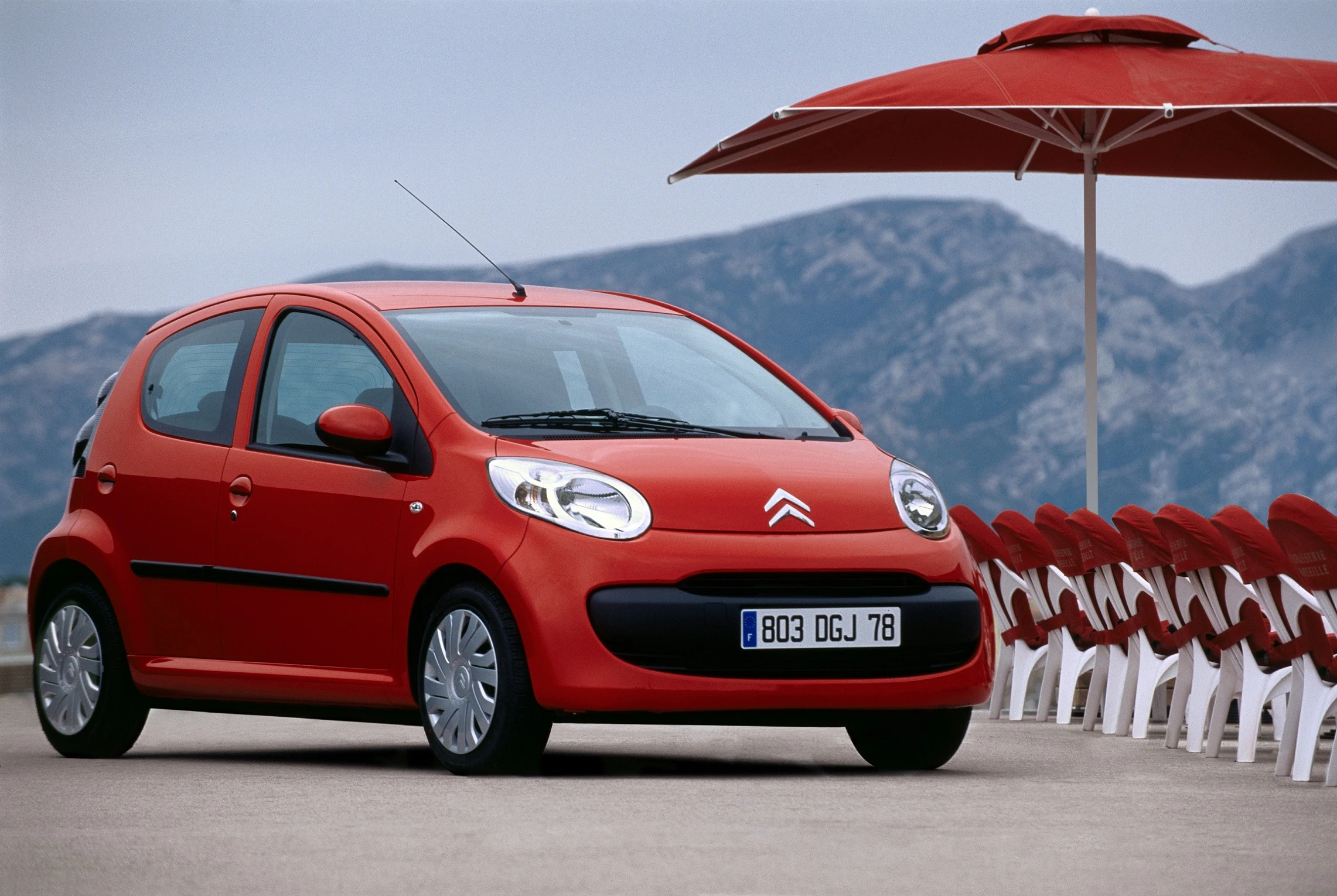 Rotes Auto auf Parkplatz mit weißen Plastikstühlen und rotem Sonnenschirm, im Hintergrund Berge und bewölkter Himmel.