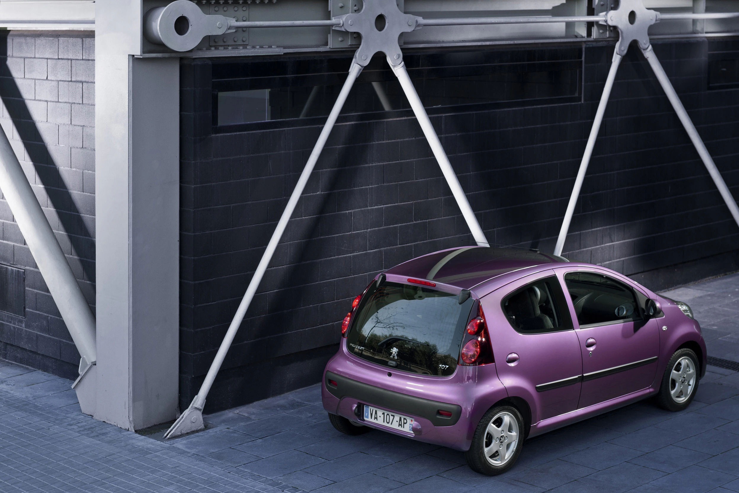 Lila Auto parkt vor einem modernen Gebäude mit sichtbaren Stützstrukturen, Wand aus dunklen Ziegeln, graue Fensterrahmen.
