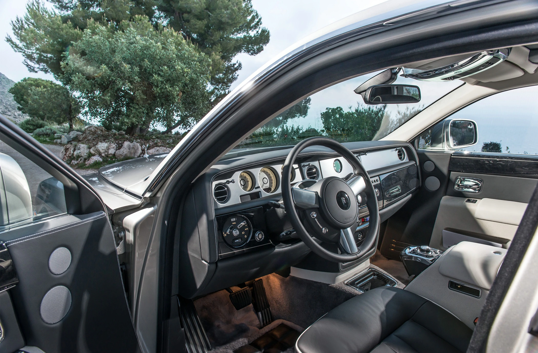 Innenraum eines luxuriösen Autos mit Lederlenkrad und eleganter Armaturentafel, Blick nach draußen auf Bäume und bewölkten Himmel.