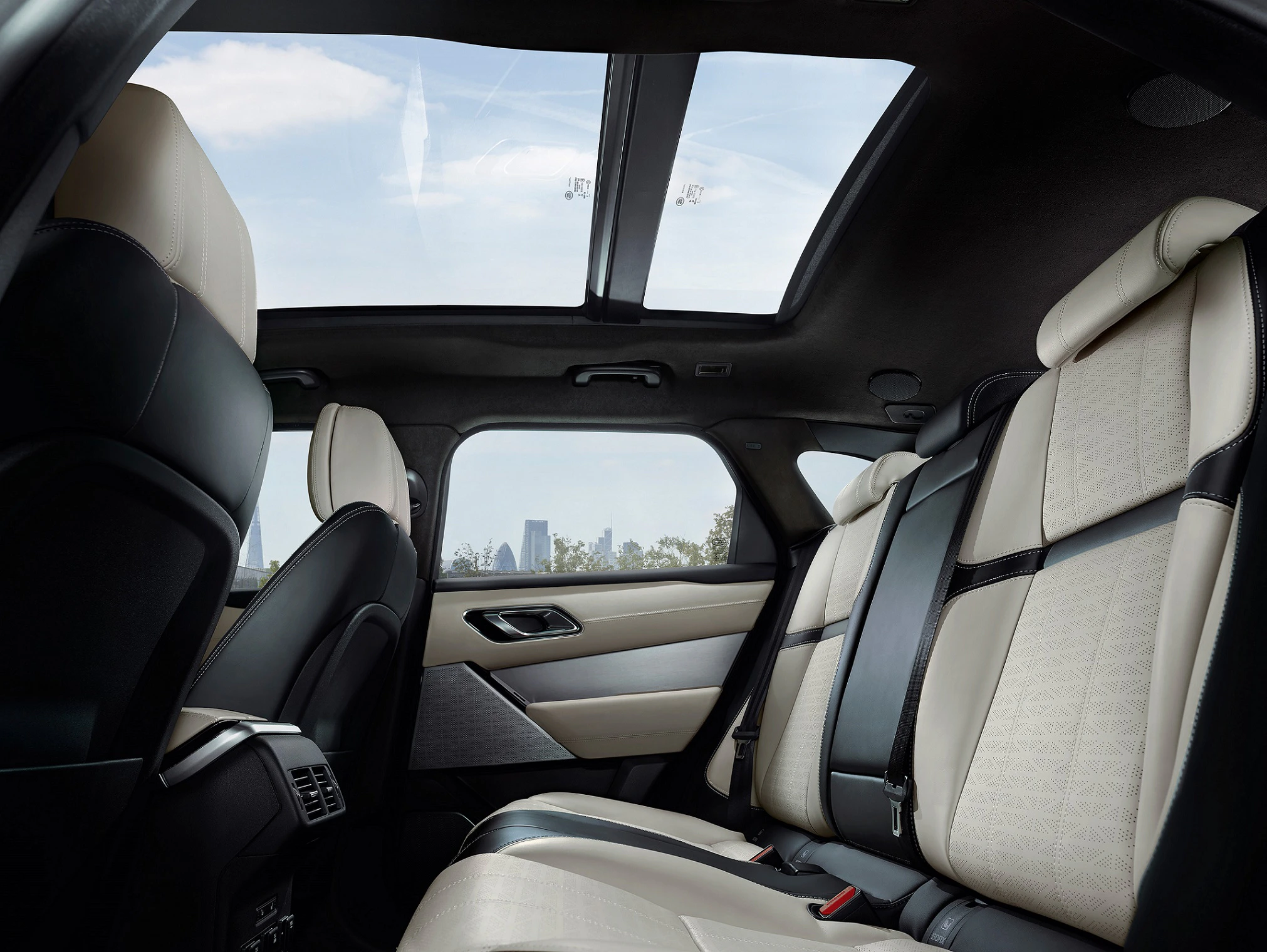 Innenraum eines Autos mit Panoramaglasdach. Helle Ledersitze und Blick auf den Himmel und eine Stadtlandschaft im Hintergrund.