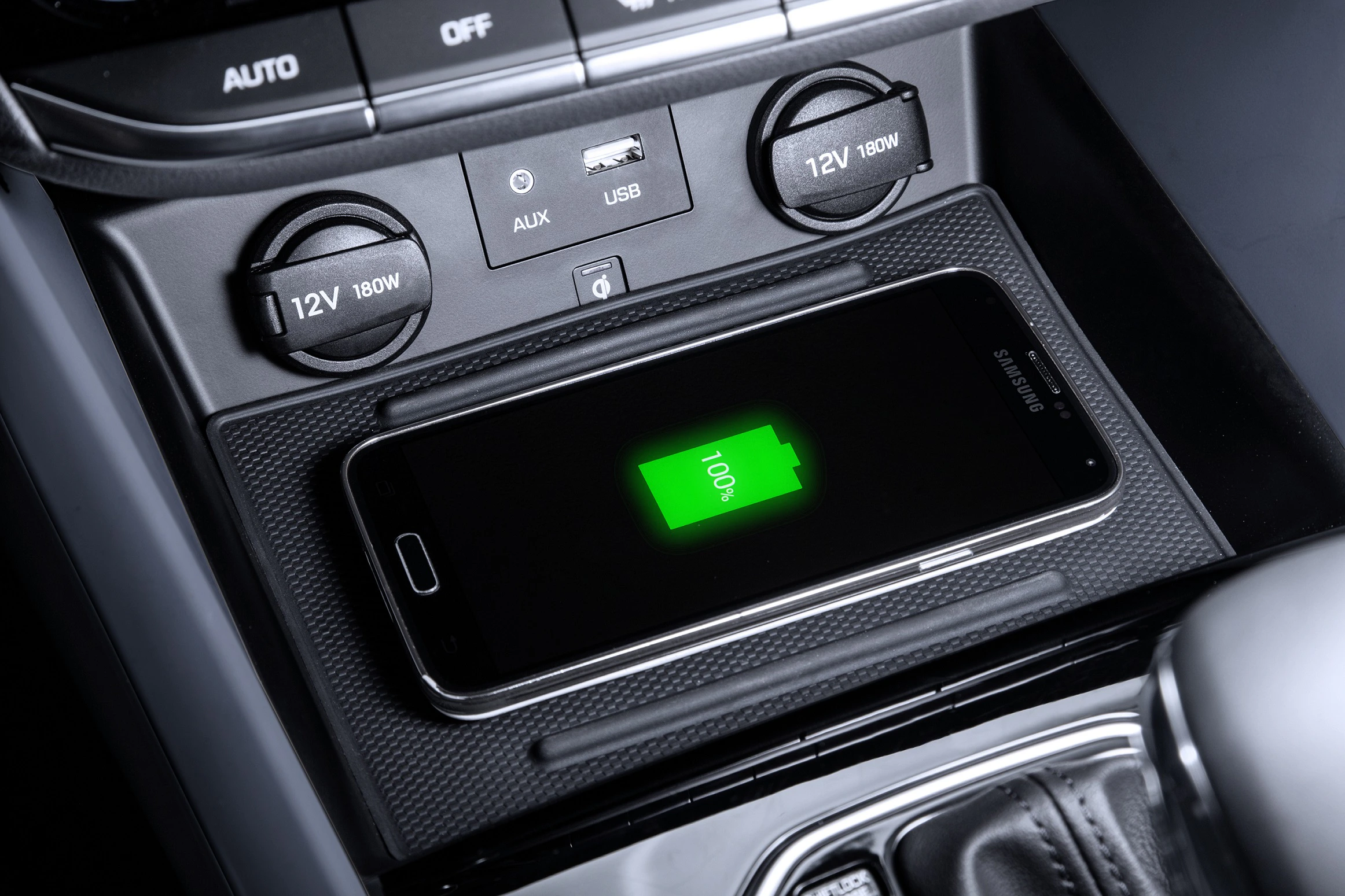 Smartphone lädt kabellos in einer Auto-Konsole. Anzeige weist auf 100% Ladestand hin. Umgebung zeigt USB- und AUX-Anschlüsse.