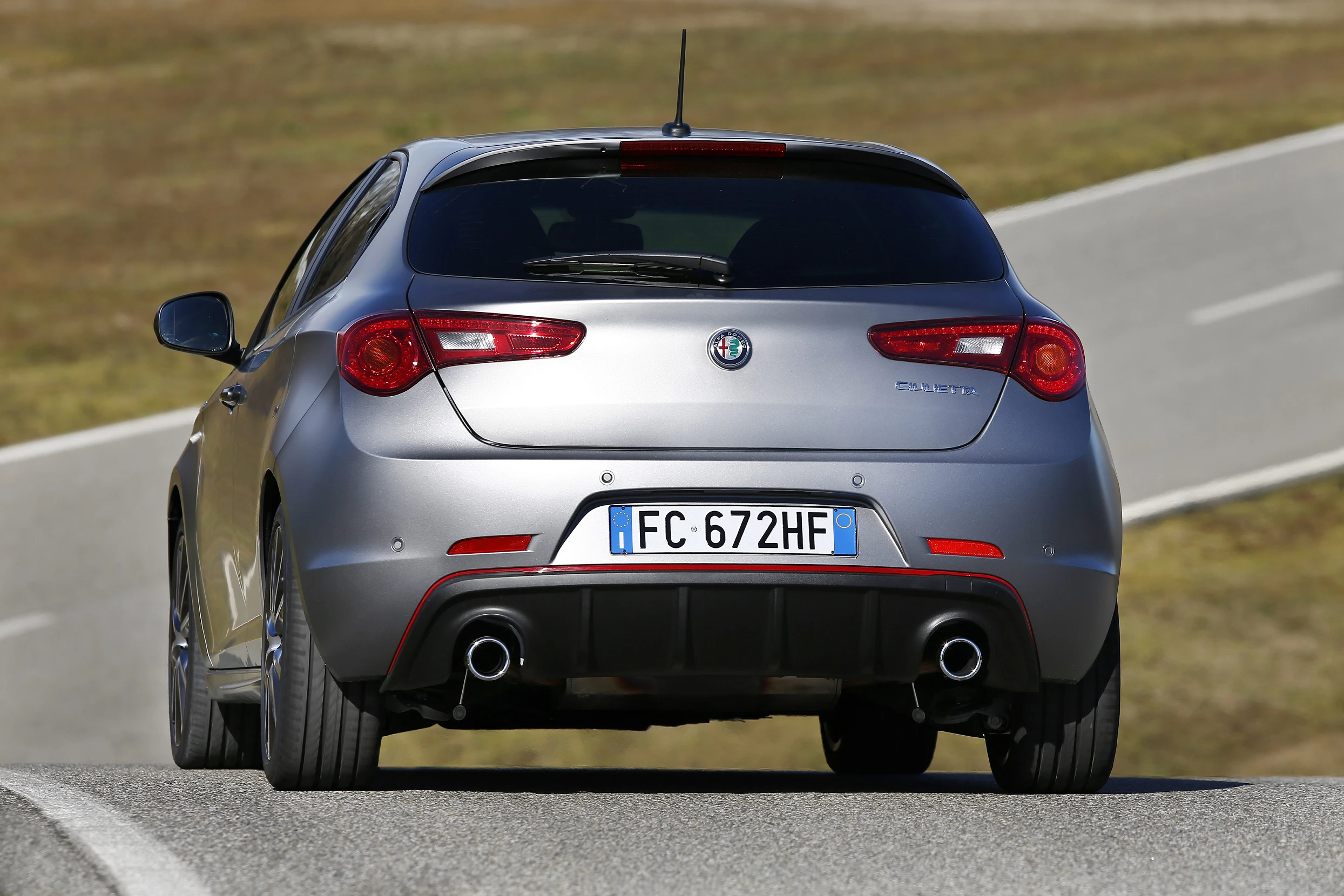 Silberner Alfa Romeo Giulietta fährt auf einer kurvigen Straße, Rückansicht sichtbar mit italienischem Kennzeichen.