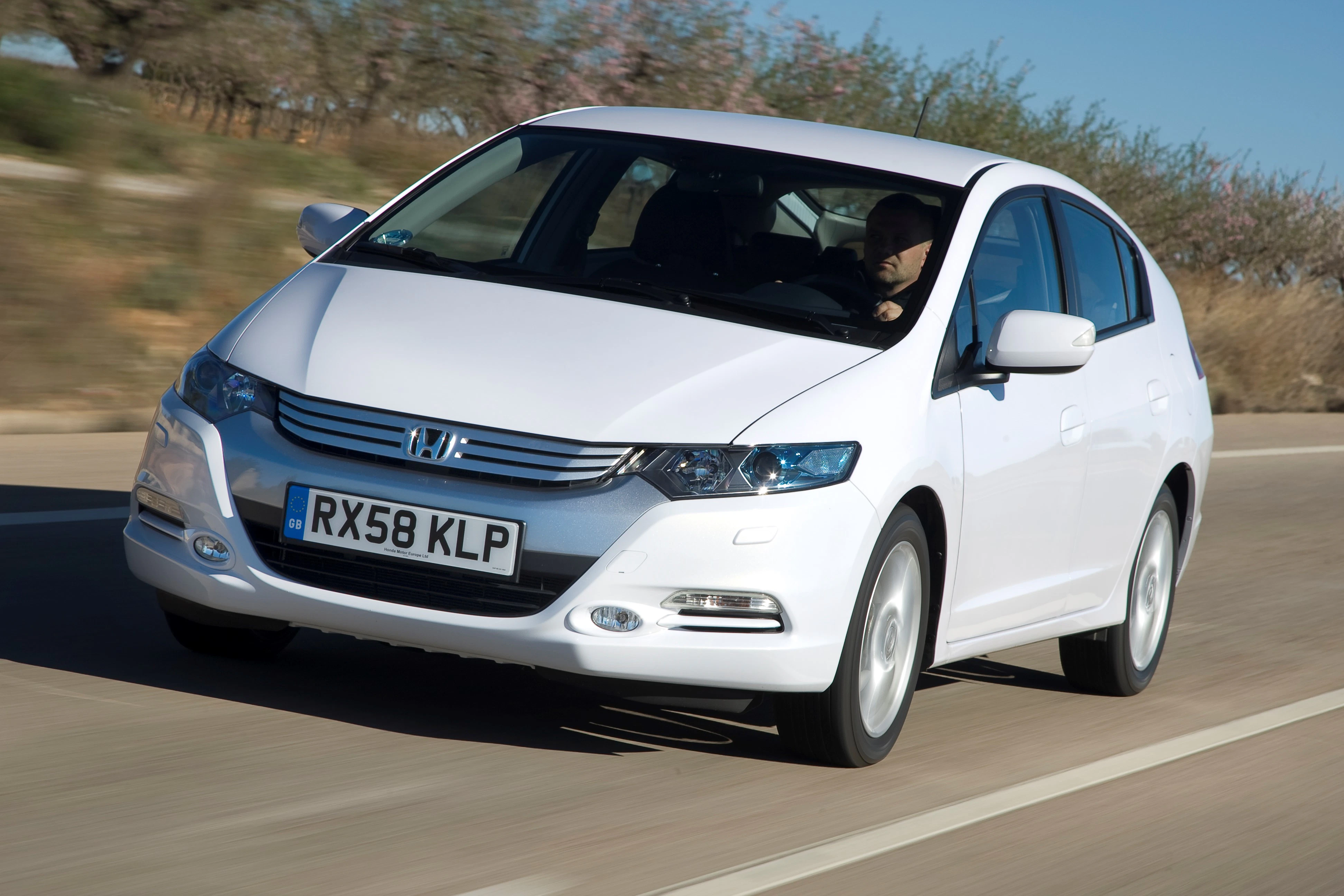 Weißer Honda Insight auf einer Landstraße, Fahrer am Steuer, Bäume im Hintergrund, bewölkter Himmel.