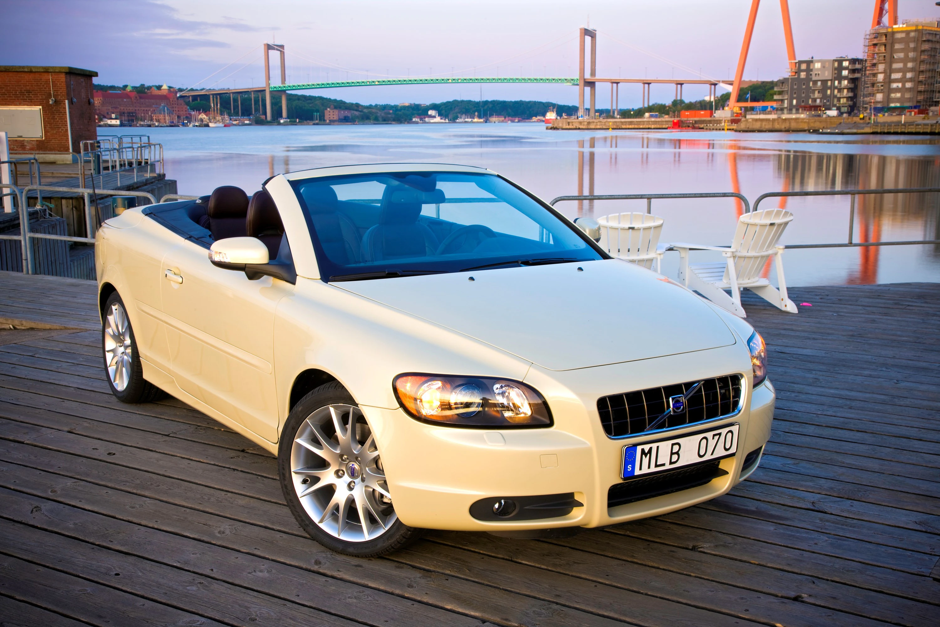 Ein weißes Cabrio steht auf einem Steg am Wasser, mit einer Brücke im Hintergrund bei Sonnenuntergang.