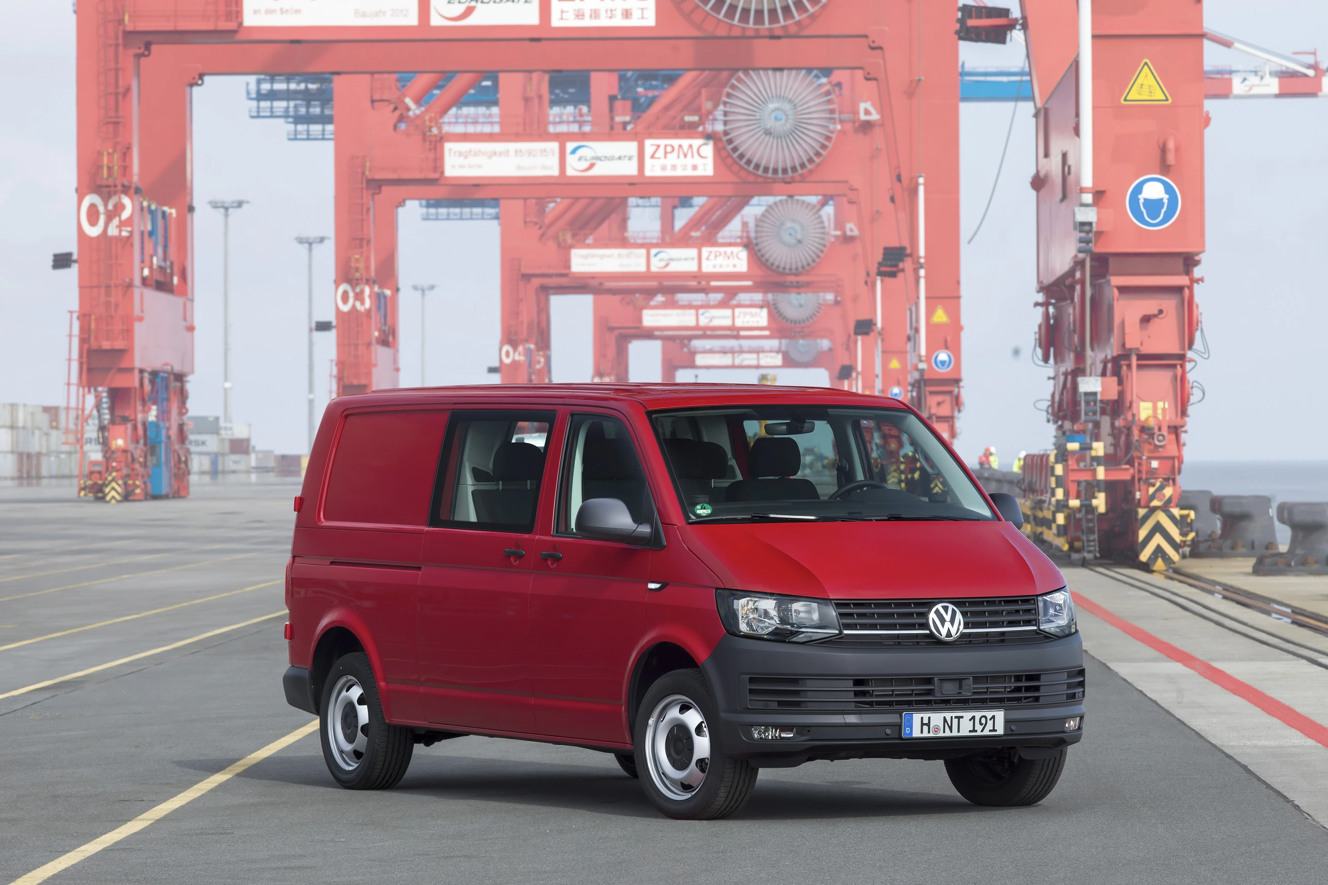 Roter Volkswagen Transporter steht auf einem Industriehafen mit roten Containerkränen im Hintergrund.