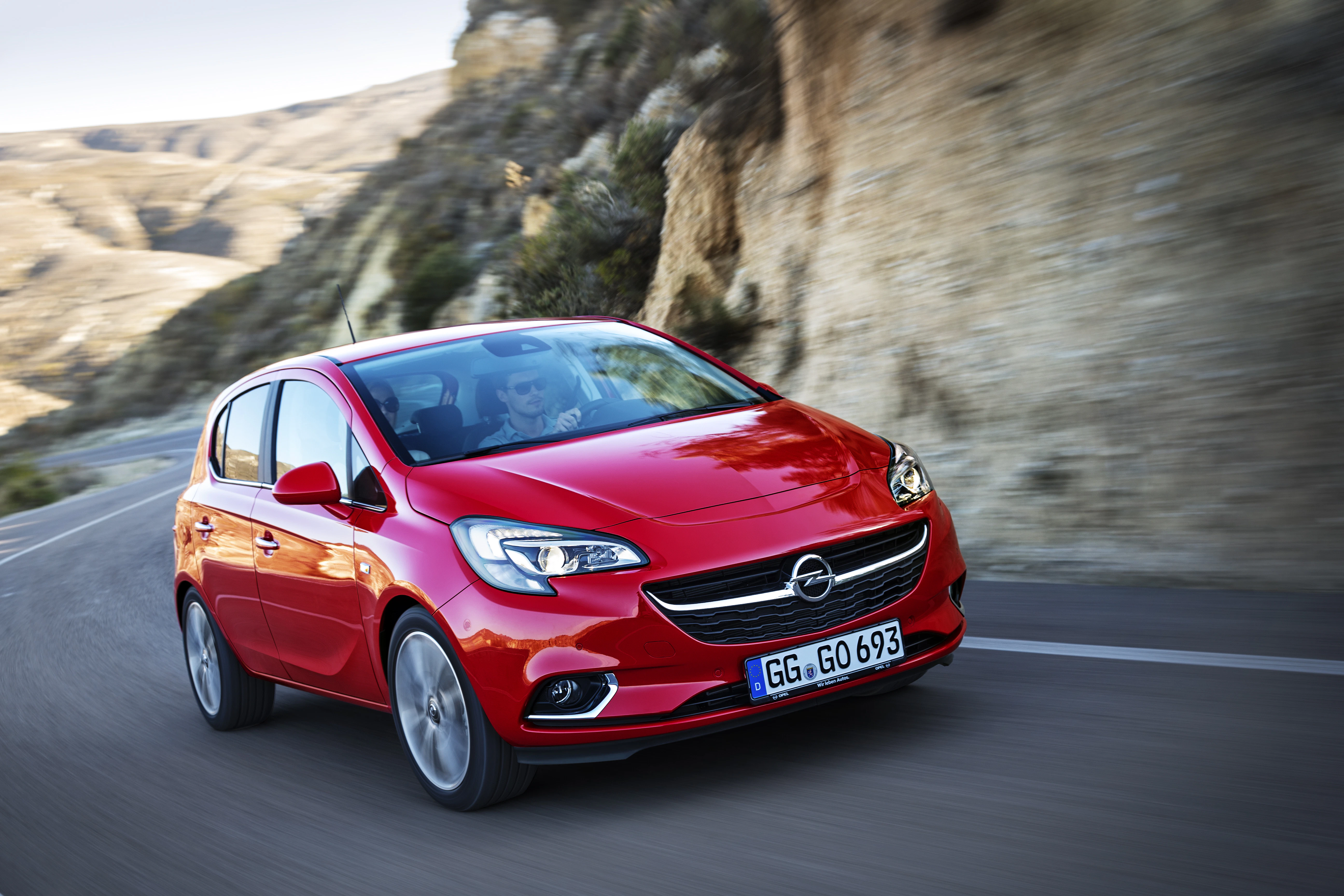 Roter Opel Corsa fährt dynamisch auf einer kurvigen Bergstraße mit felsiger Landschaft im Hintergrund bei klarem Wetter.