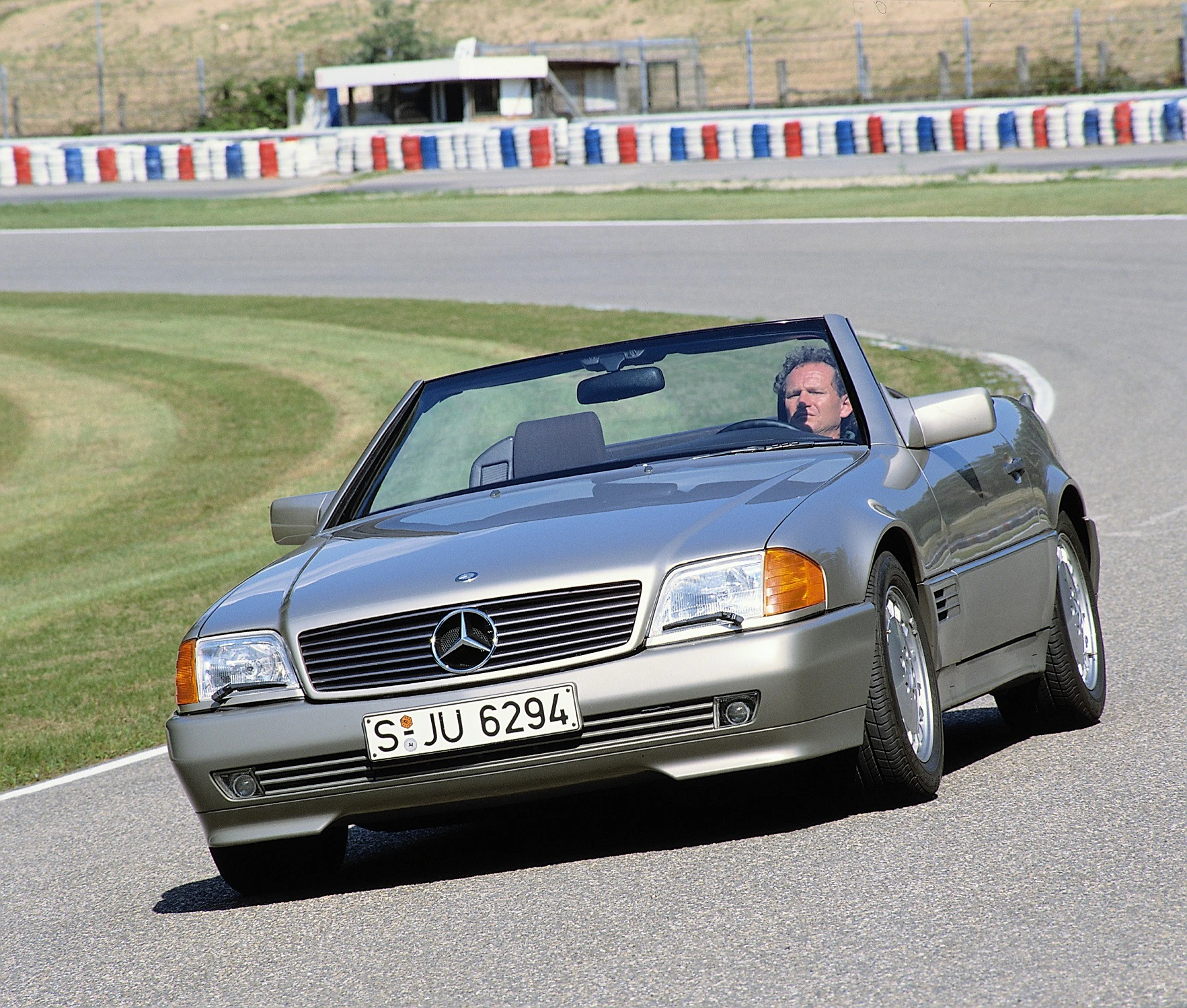 Graues Mercedes-Cabrio fährt auf Rennstrecke. Einzelner Fahrer am Steuer, keine weiteren Fahrzeuge in Sicht, klare Wetterbedingungen.