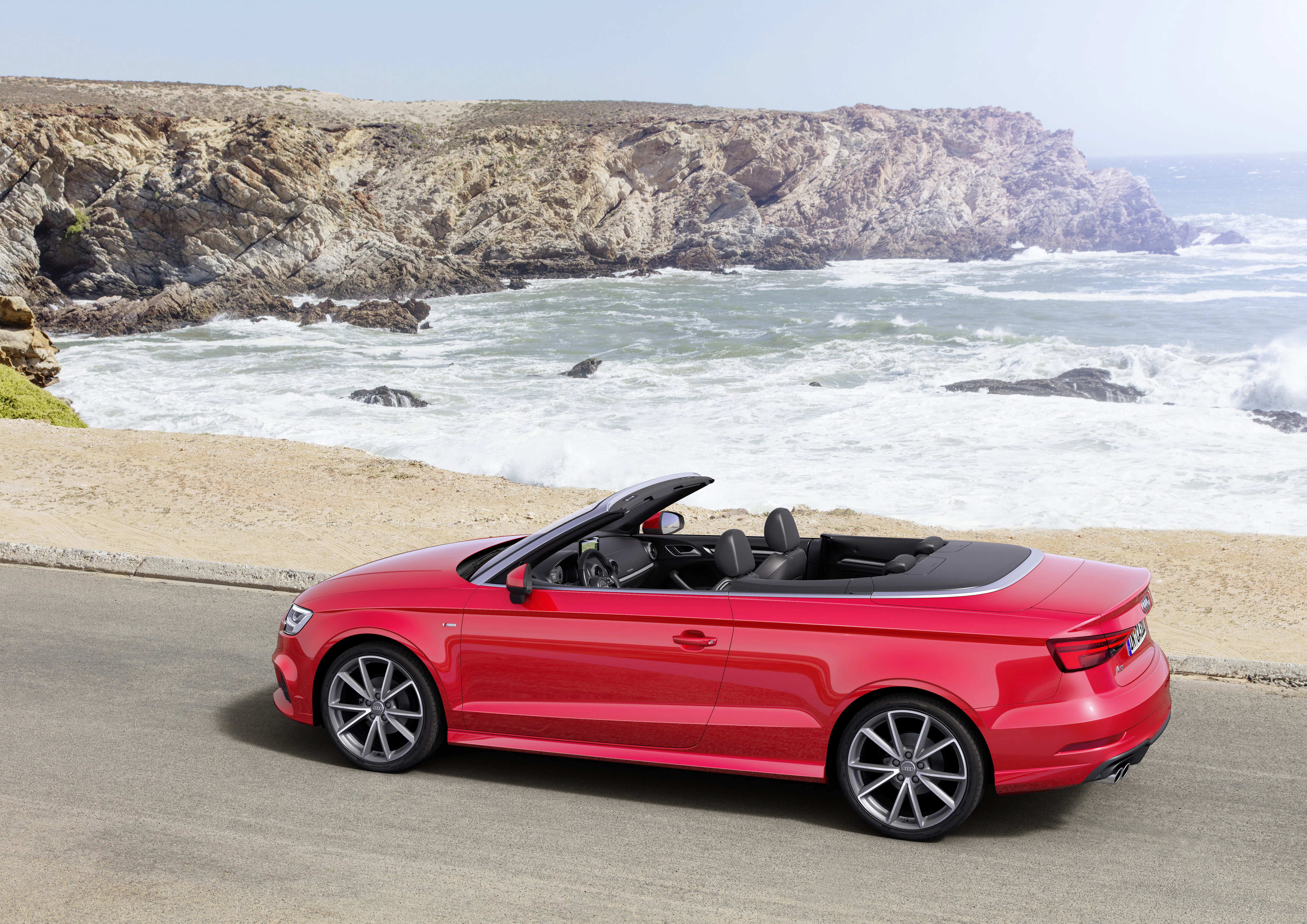 Rotes Cabrio auf Küstenstraße vor felsiger Meereskulisse bei sonnigem Wetter.