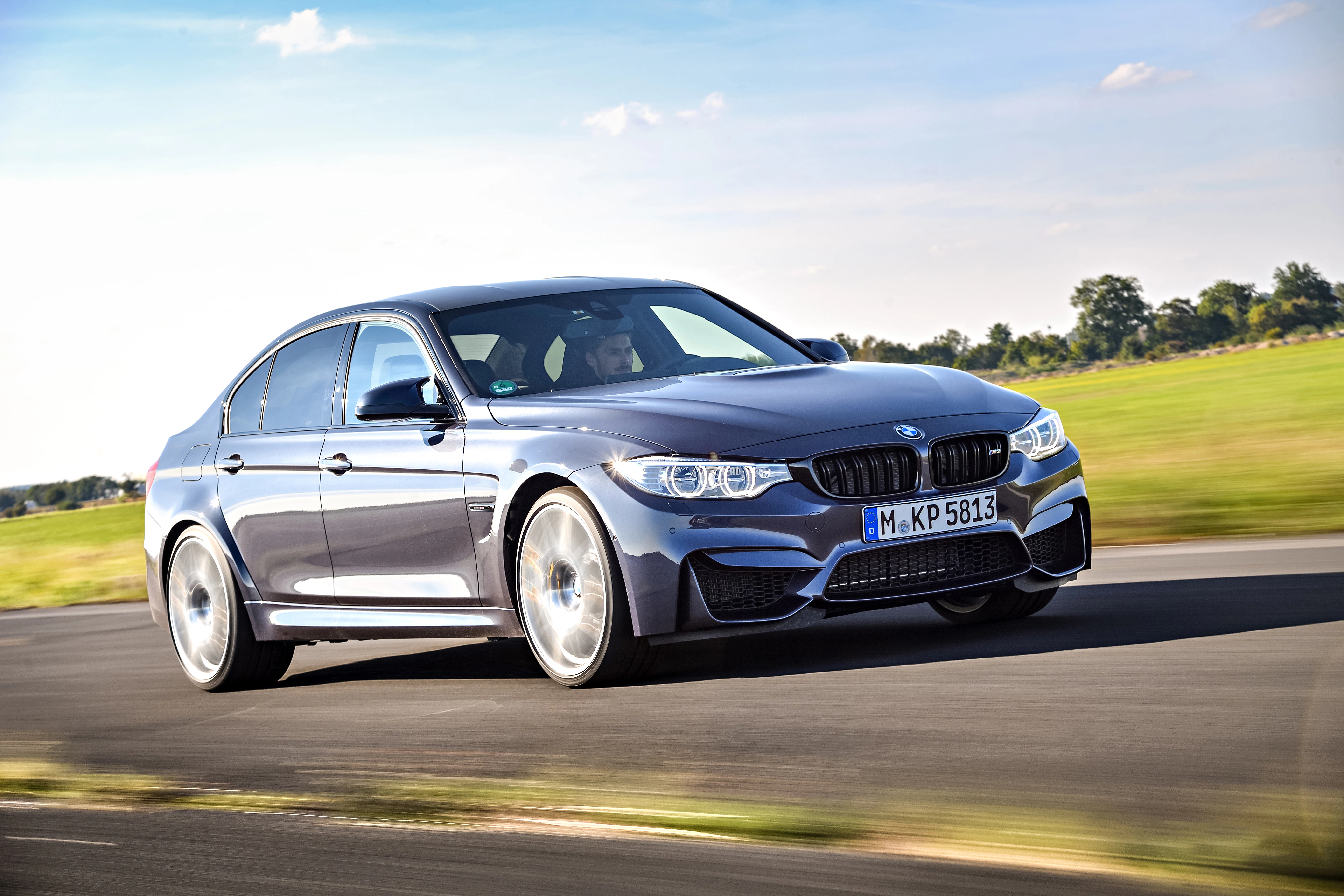 Grauer BMW auf einer Landstraße, im Hintergrund eine grüne Wiese und blauer Himmel. Fahrzeug bewegt sich dynamisch nach rechts.