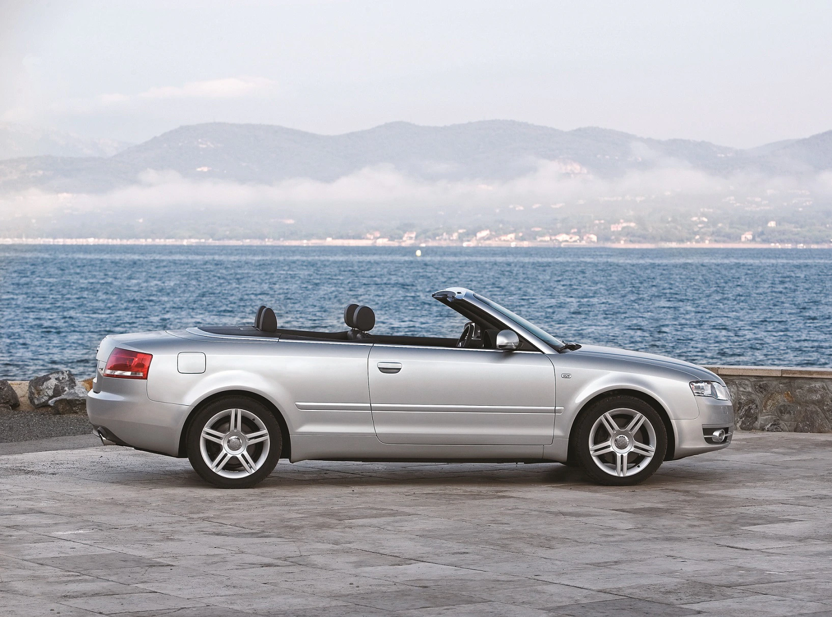 Silbernes Cabriolet geparkt am Ufer mit Blick auf Wasser und Berge im Hintergrund.
