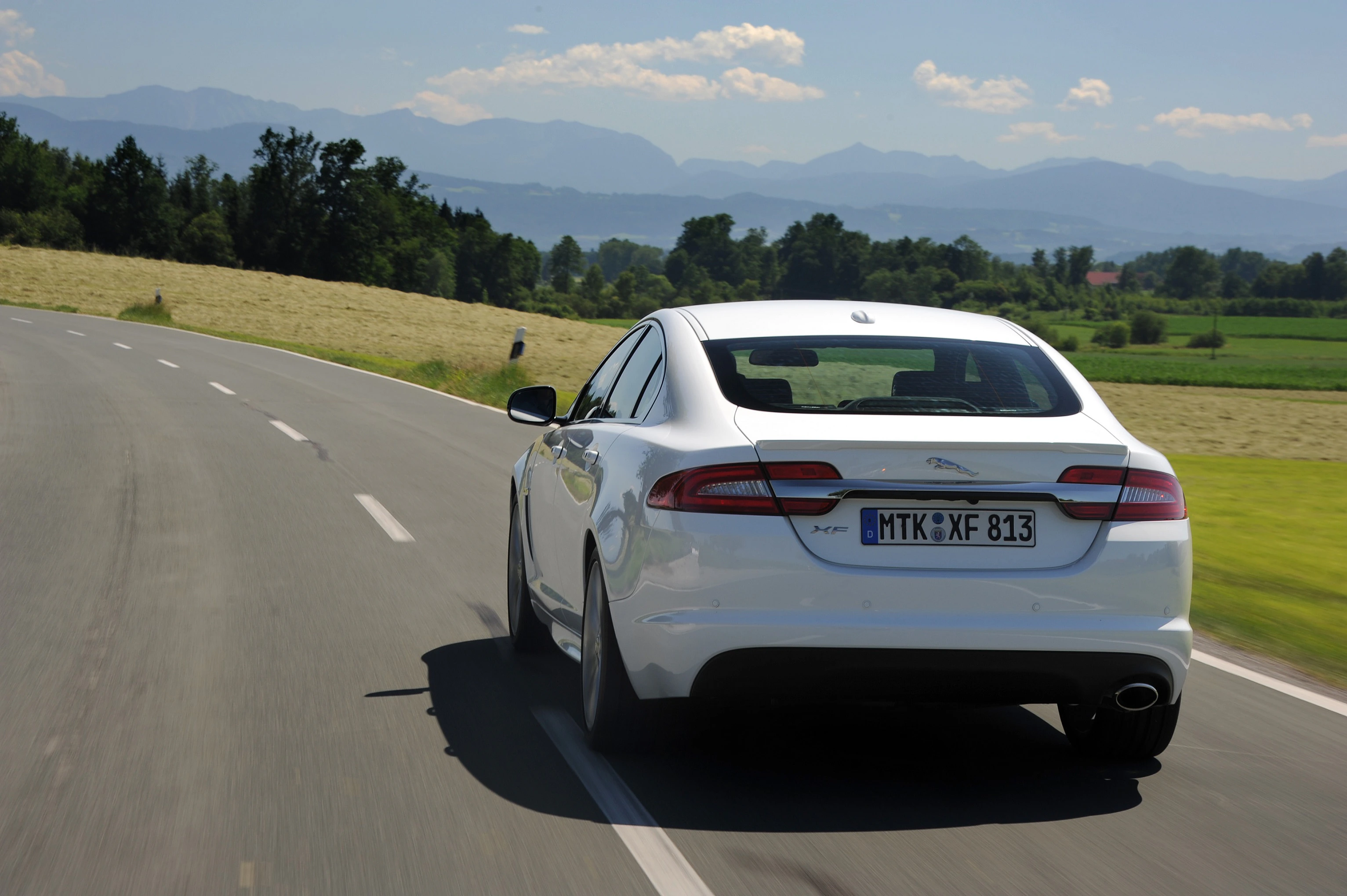 Weißes Auto fährt auf einer Landstraße durch eine grüne Landschaft mit Bergen im Hintergrund unter klarem Himmel.