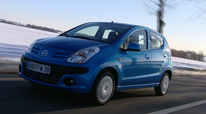Blauer Kleinwagen fährt auf winterlicher Landstraße, Schneelandschaft im Hintergrund, Baumreihe am Rand der Straße.