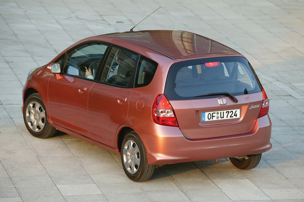 Roter Honda Jazz in seitlicher Heckansicht auf gepflastertem Untergrund, mit sichtbarem Nummernschild und geschlossenen Fenstern.