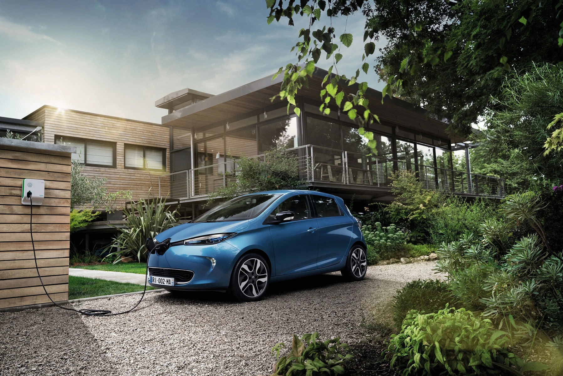 Blaues Elektroauto lädt an einer Ladestation neben einem modernen Haus im Grünen auf, umgeben von üppiger Vegetation und klarem Himmel.