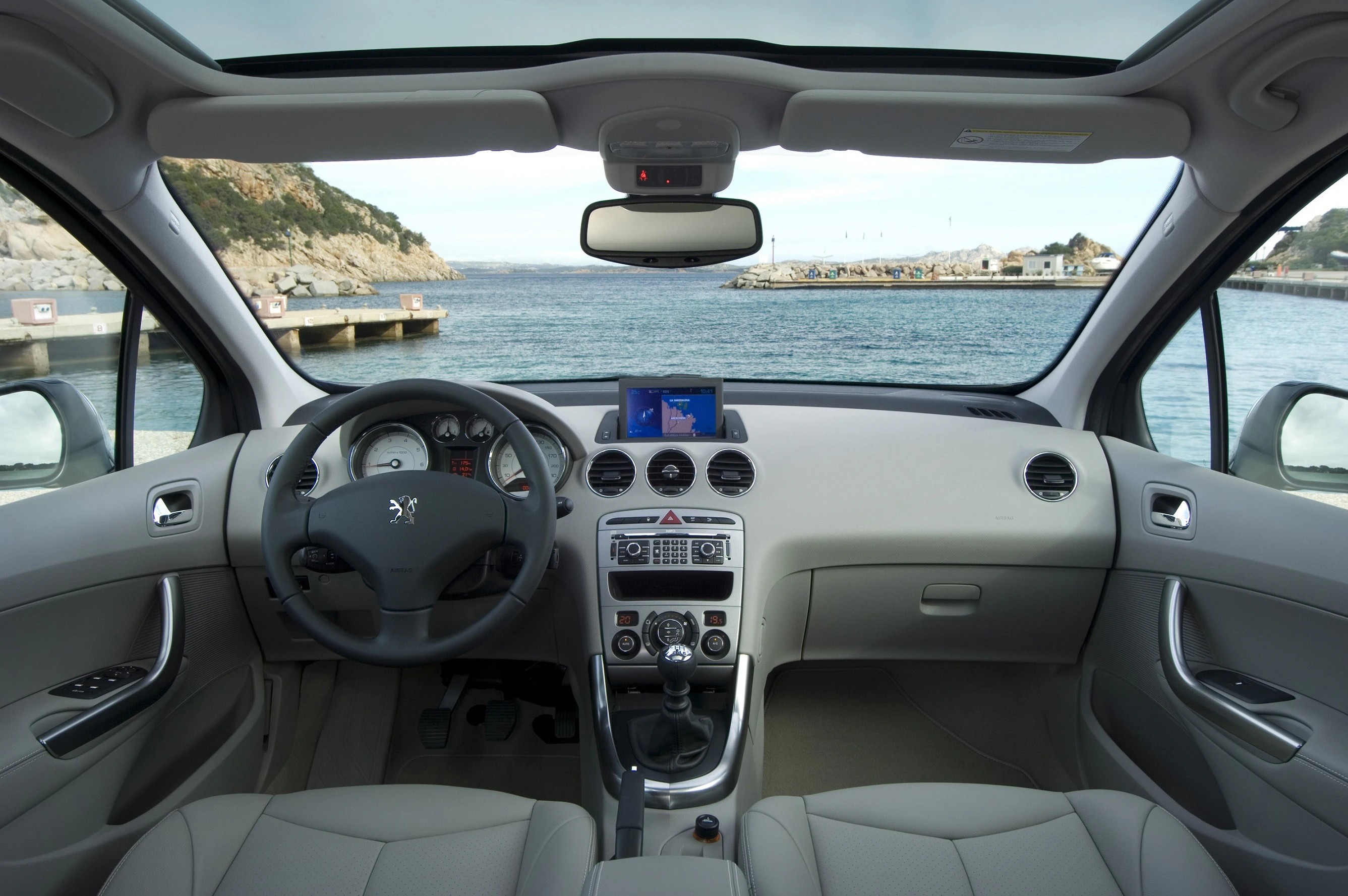 Innenraum eines Autos mit Blick durch die Windschutzscheibe auf das Meer und eine Küstenlandschaft. Lenkrad und Armaturenbrett sichtbar.