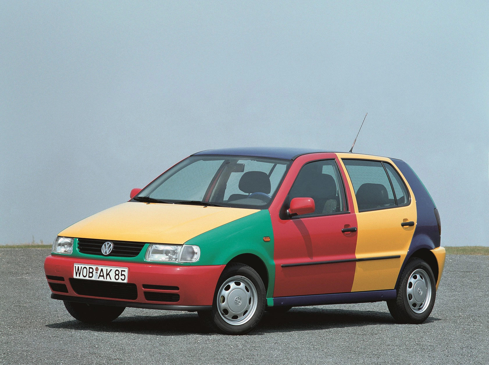 Bunter VW Polo mit vierfarbiger Lackierung in Gelb, Grün, Rot und Blau steht auf asphaltierter Fläche, mit grauem Himmel im Hintergrund.