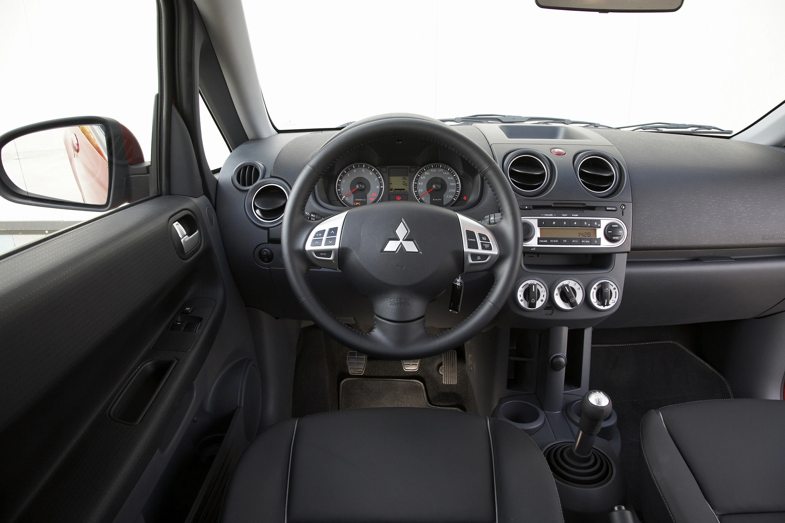 Innenraum eines Mitsubishi mit Lenkrad, Tachometer und Bedienelementen. Schlichtes Armaturenbrettdesign mit Handschaltung.