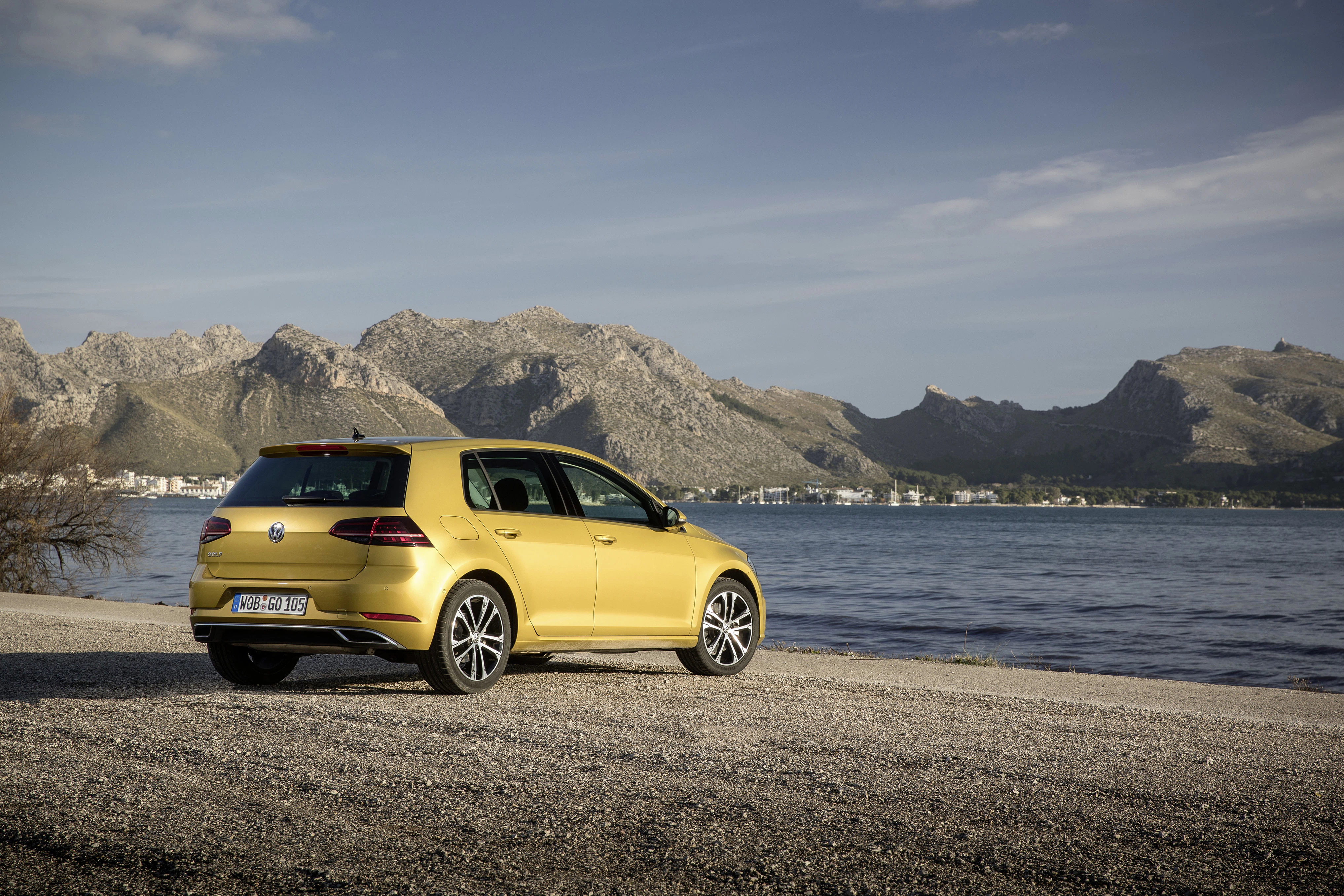 Ein gelber Volkswagen Golf parkt am Ufer eines Sees mit Bergen im Hintergrund unter klarem Himmel.