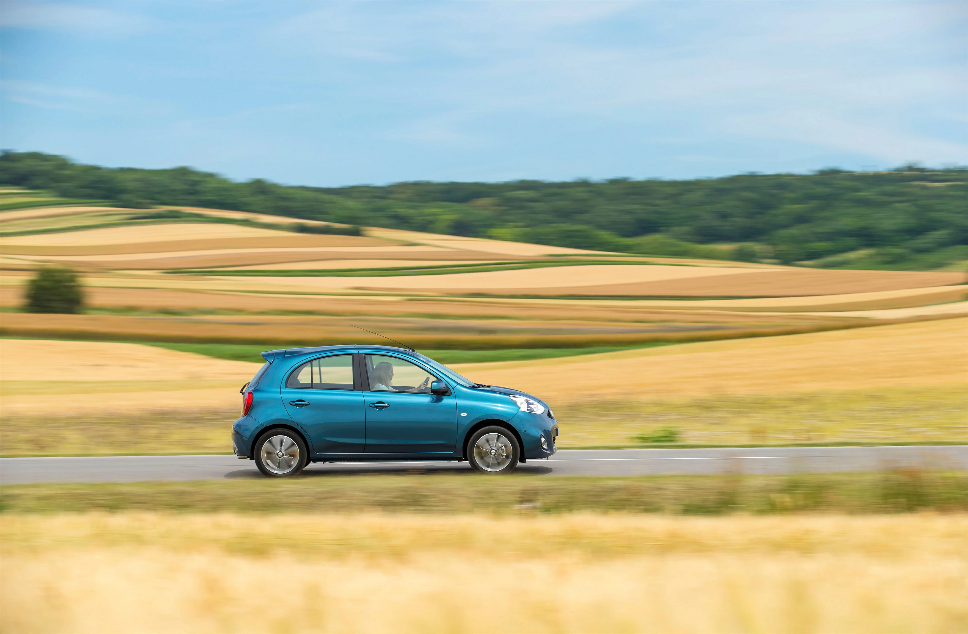 Kleines, blaues Auto fährt schnell auf einer Landstraße durch eine malerische, hügelige Landschaft mit Feldern und Wäldern unter blauem Himmel.