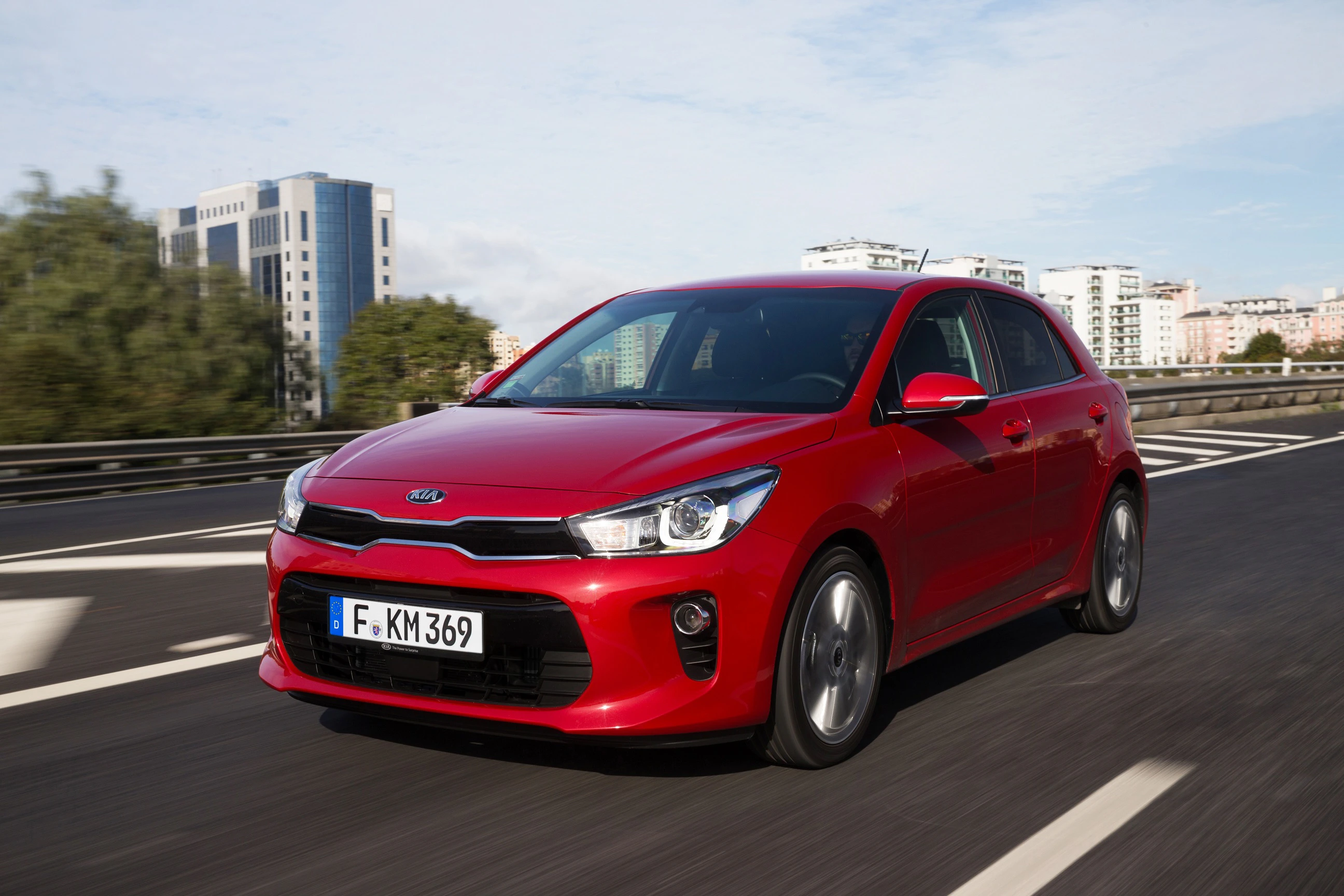 Roter Kia Rio fährt auf einer städtischen Autobahn bei Tageslicht. Im Hintergrund sind moderne Gebäude sichtbar.