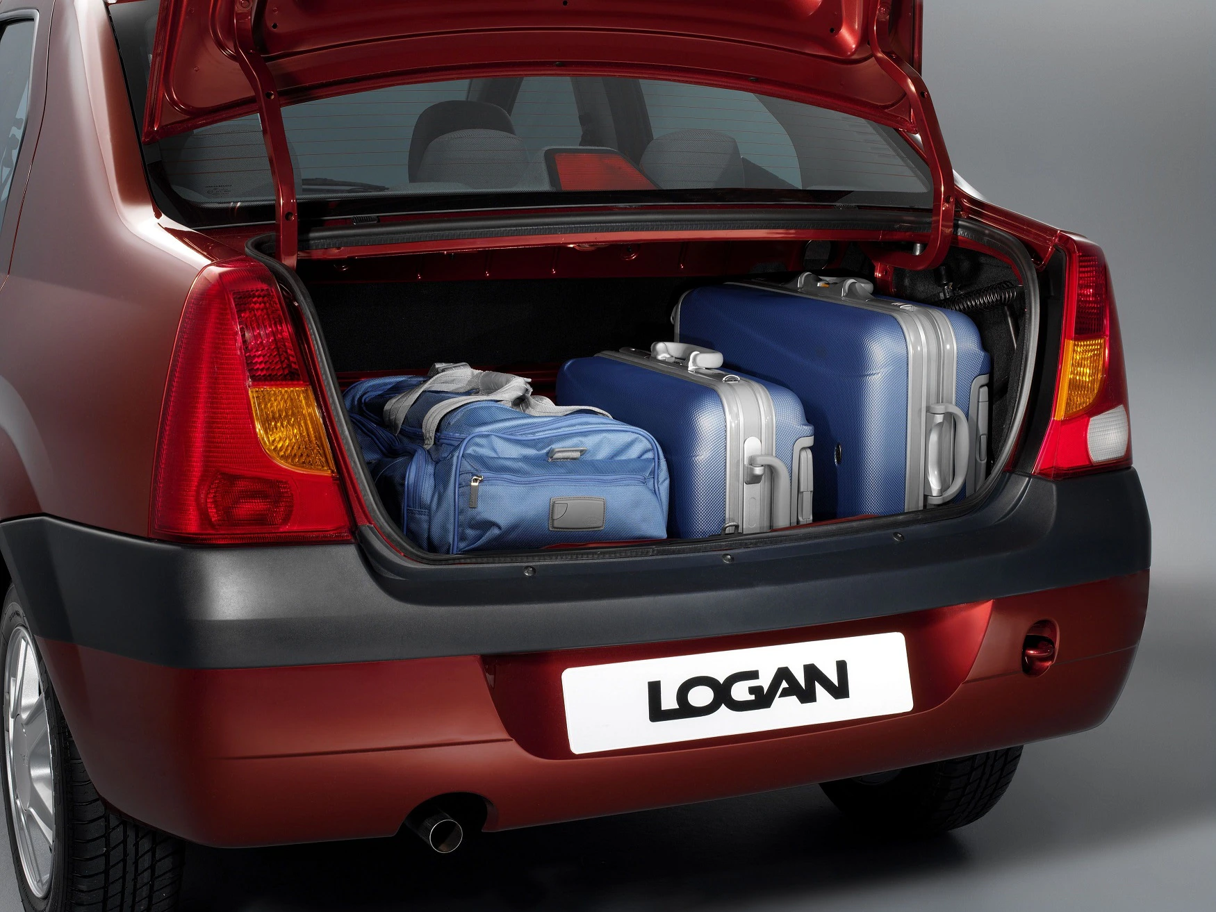 Auto mit offenem Kofferraum, der mehrere Reisekoffer zeigt. Zwei blaue und ein silberner Koffer sind sichtbar. Nummernschild: "LOGAN".