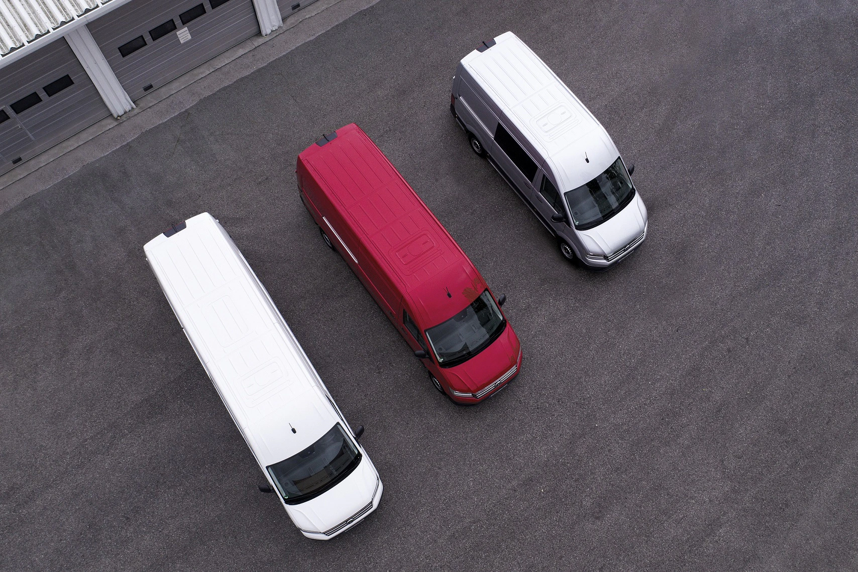 Drei Transporter von oben abgebildet: links weiß, in der Mitte rot, rechts grau, auf einem asphaltierten Platz vor Garagentoren.