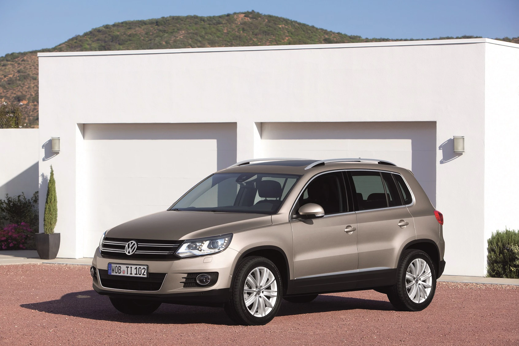 Ein brauner Volkswagen Tiguan steht vor einem modernen weißen Gebäude mit zwei Garagen und einer Berglandschaft im Hintergrund.