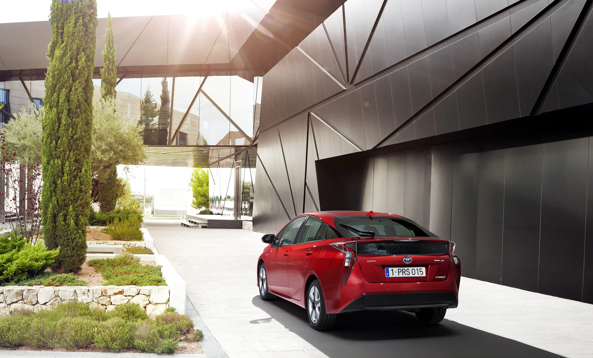Rotes Auto in moderner, futuristischer Umgebung mit geometrischen Glasgebäuden. Sonnenlicht fällt von links ein.
