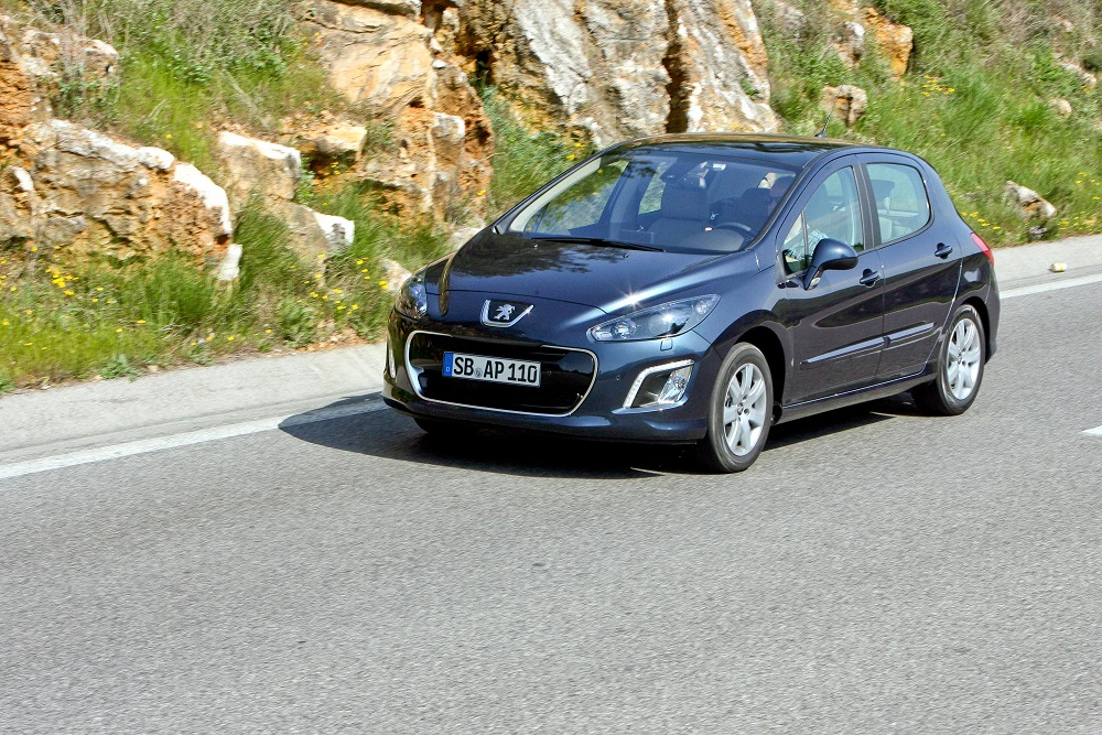 Dunkelblauer Peugeot fährt auf einer Landstraße entlang einer felsigen Landschaft. Klarer Himmel und sonniges Wetter.