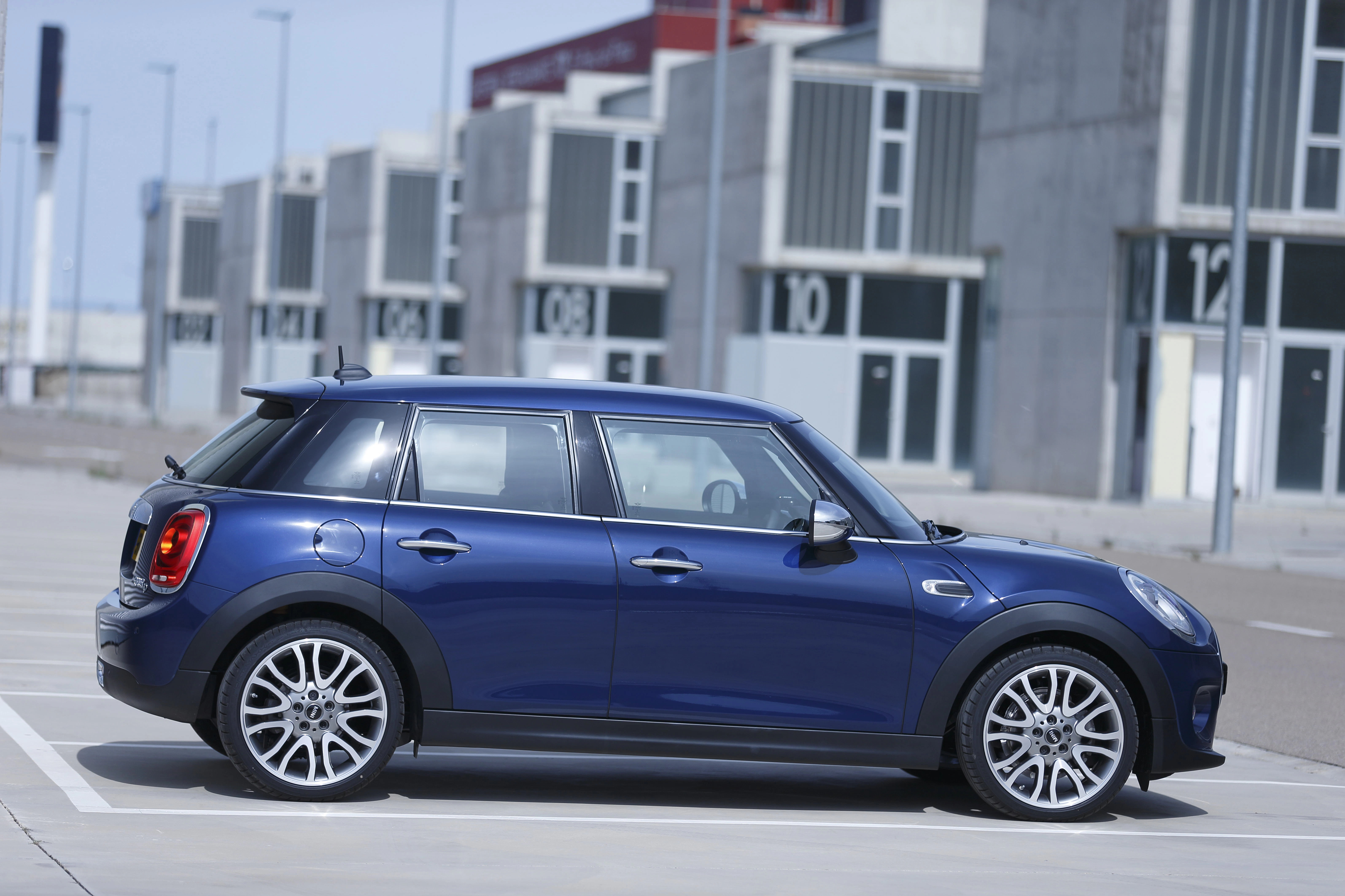 Blauer Mini Cooper steht seitlich auf einem Parkplatz, moderne Gebäude im Hintergrund.
