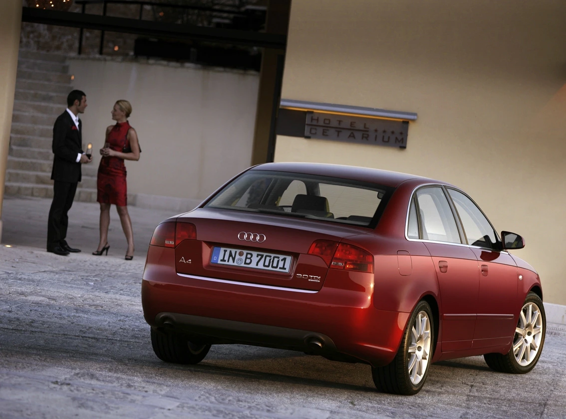 Ein roter Audi A4 steht vor einem Hotel. Im Hintergrund unterhalten sich ein Mann im Anzug und eine Frau im roten Kleid.