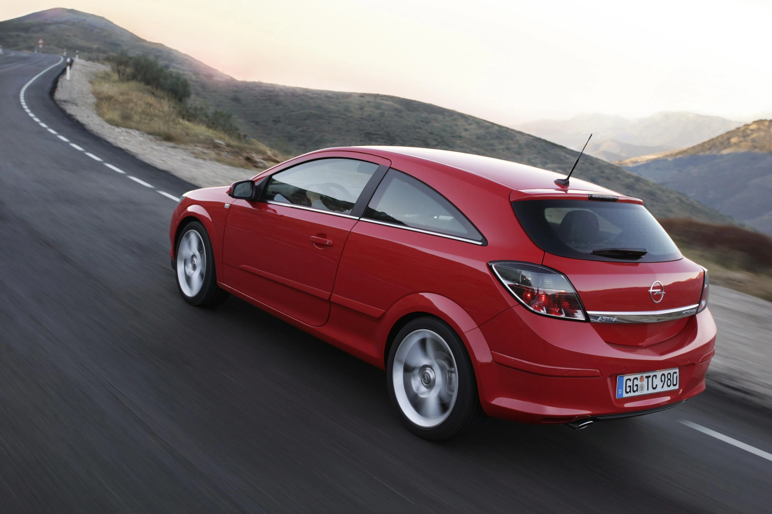 Roter Opel dreitürig fährt auf einer kurvigen Landstraße in hügeliger Landschaft bei Sonnenuntergang.