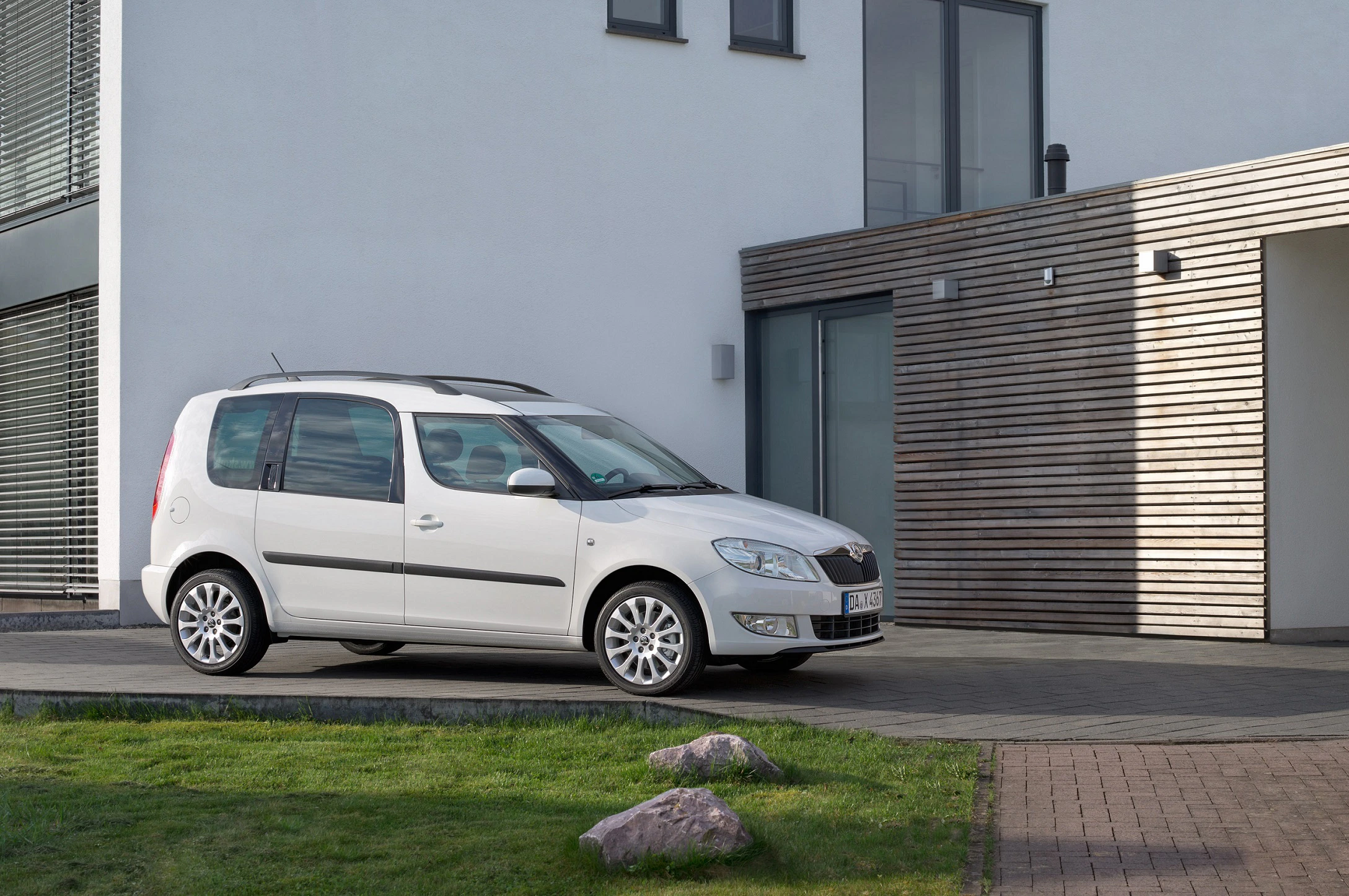 Weißer Minivan parkt vor einem modernen Haus mit Holzverkleidung und großen Fenstern. Im Vordergrund ist eine grüne Rasenfläche sichtbar.