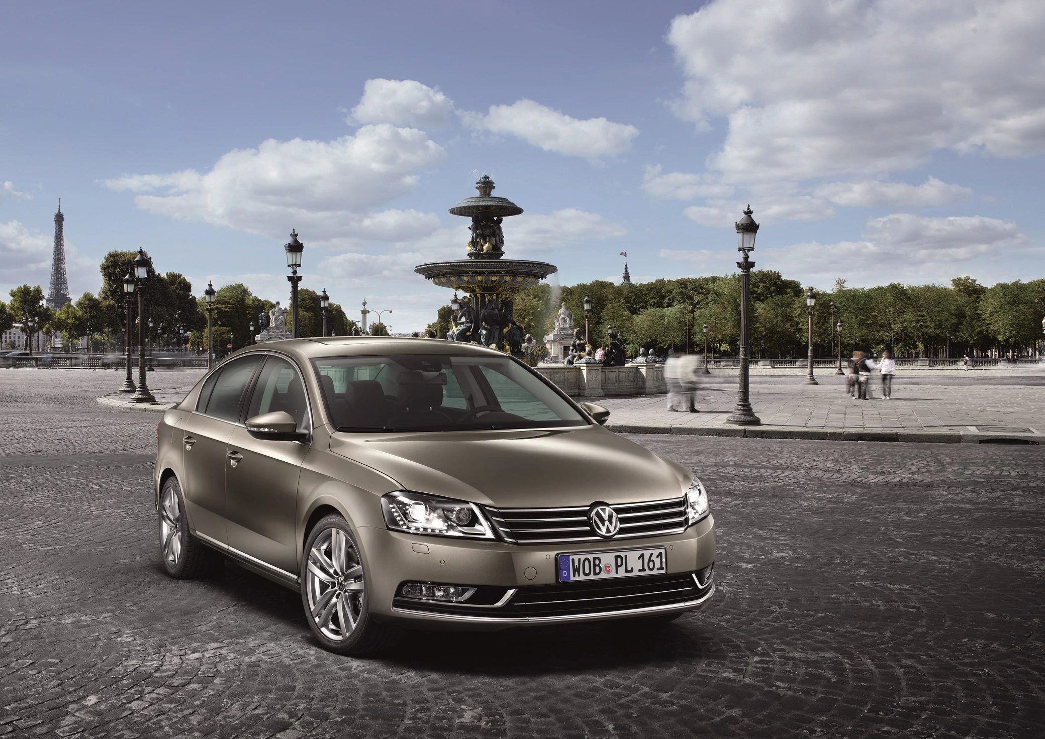 Volkswagen Passat auf einem gepflasterten Platz mit einem Brunnen im Hintergrund, umgeben von Bäumen und Laternen bei bewölktem Himmel.
