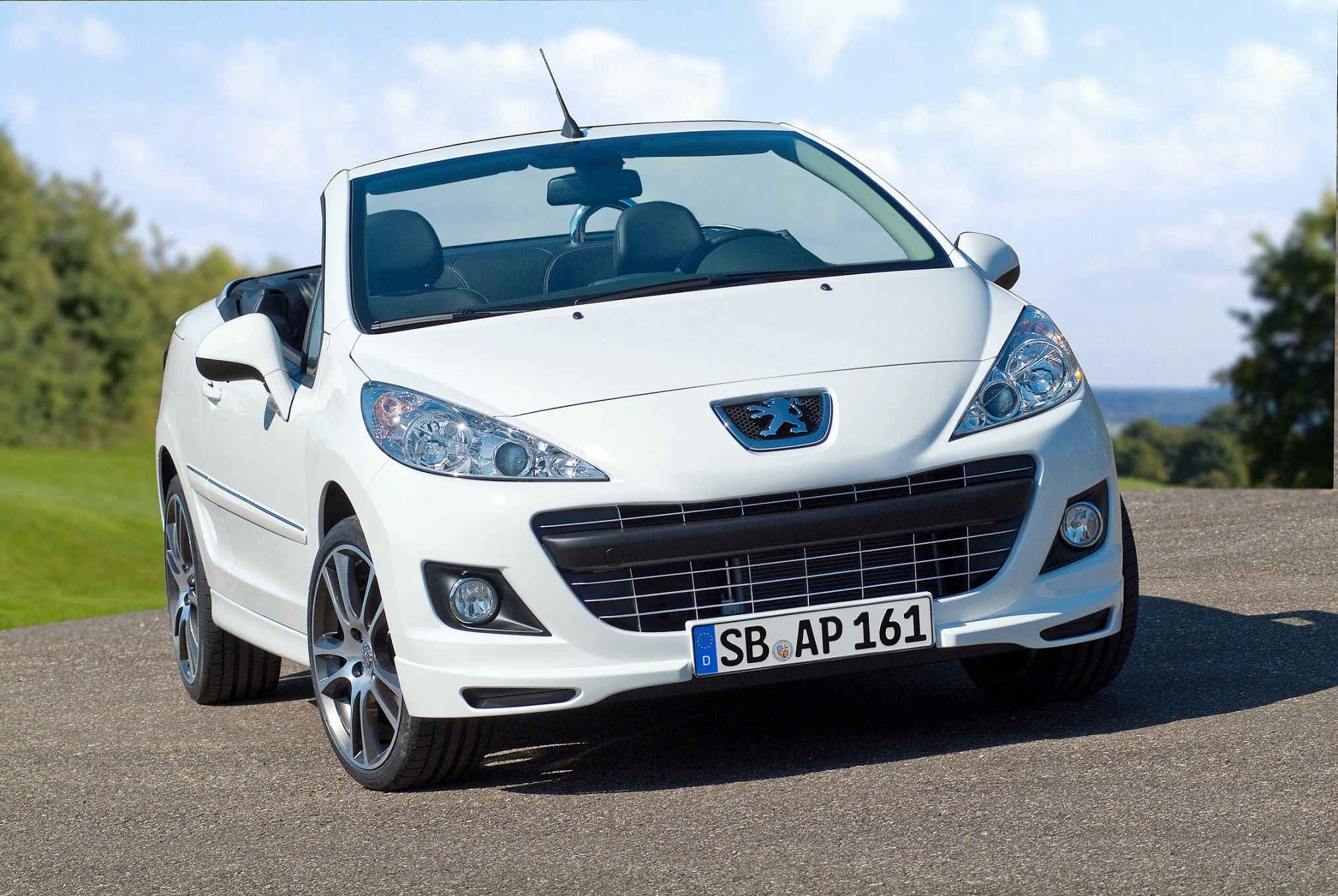 Weißes Cabriolet, Peugeot, parkt auf einer Asphaltstraße vor grünem Hintergrund mit blauem Himmel. Nummernschild: SB AP 161.