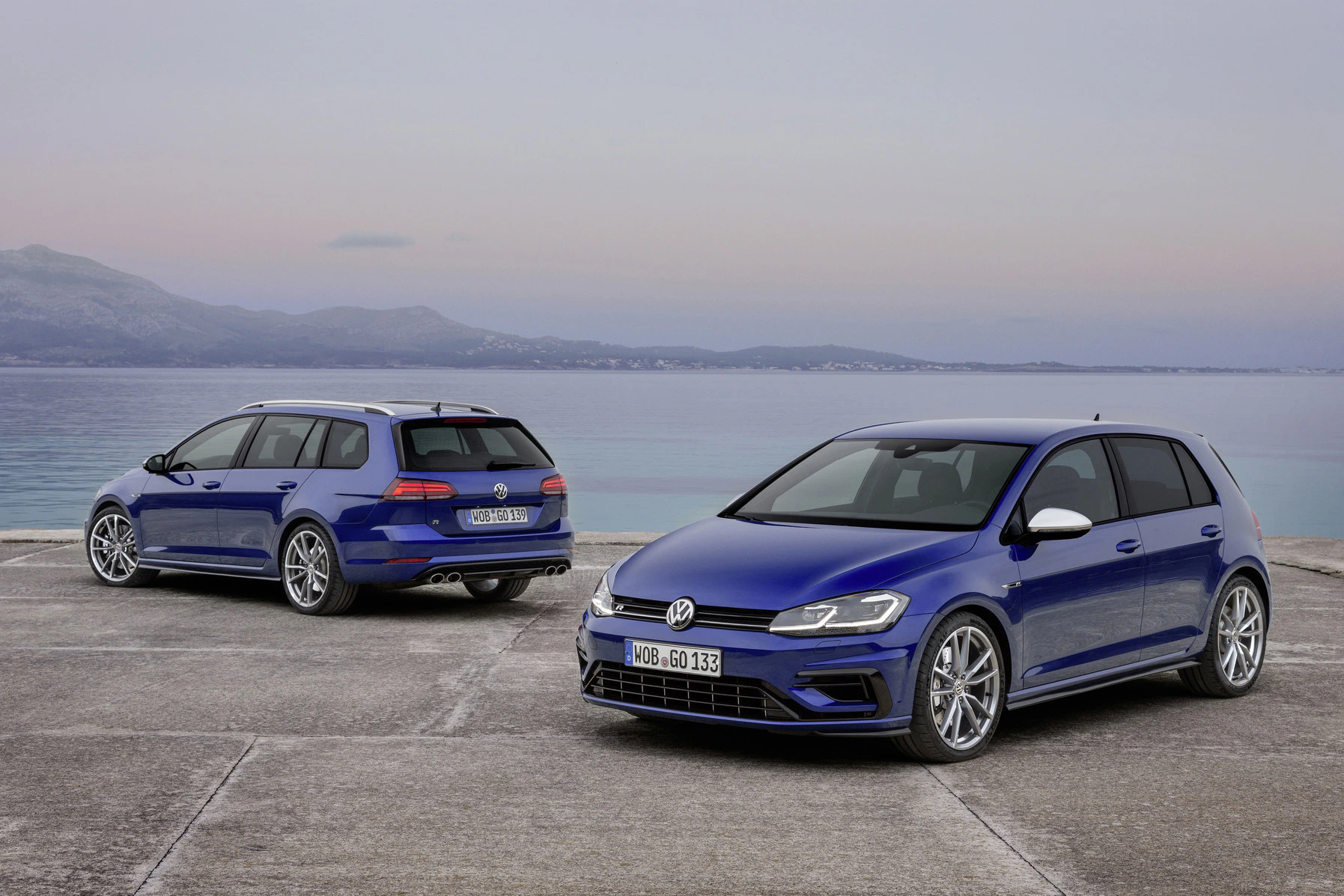 Zwei blaue Volkswagen-Autos parken auf einem Betonplatz am Meer. Der Himmel ist leicht bewölkt, und es sind Berge im Hintergrund zu sehen.