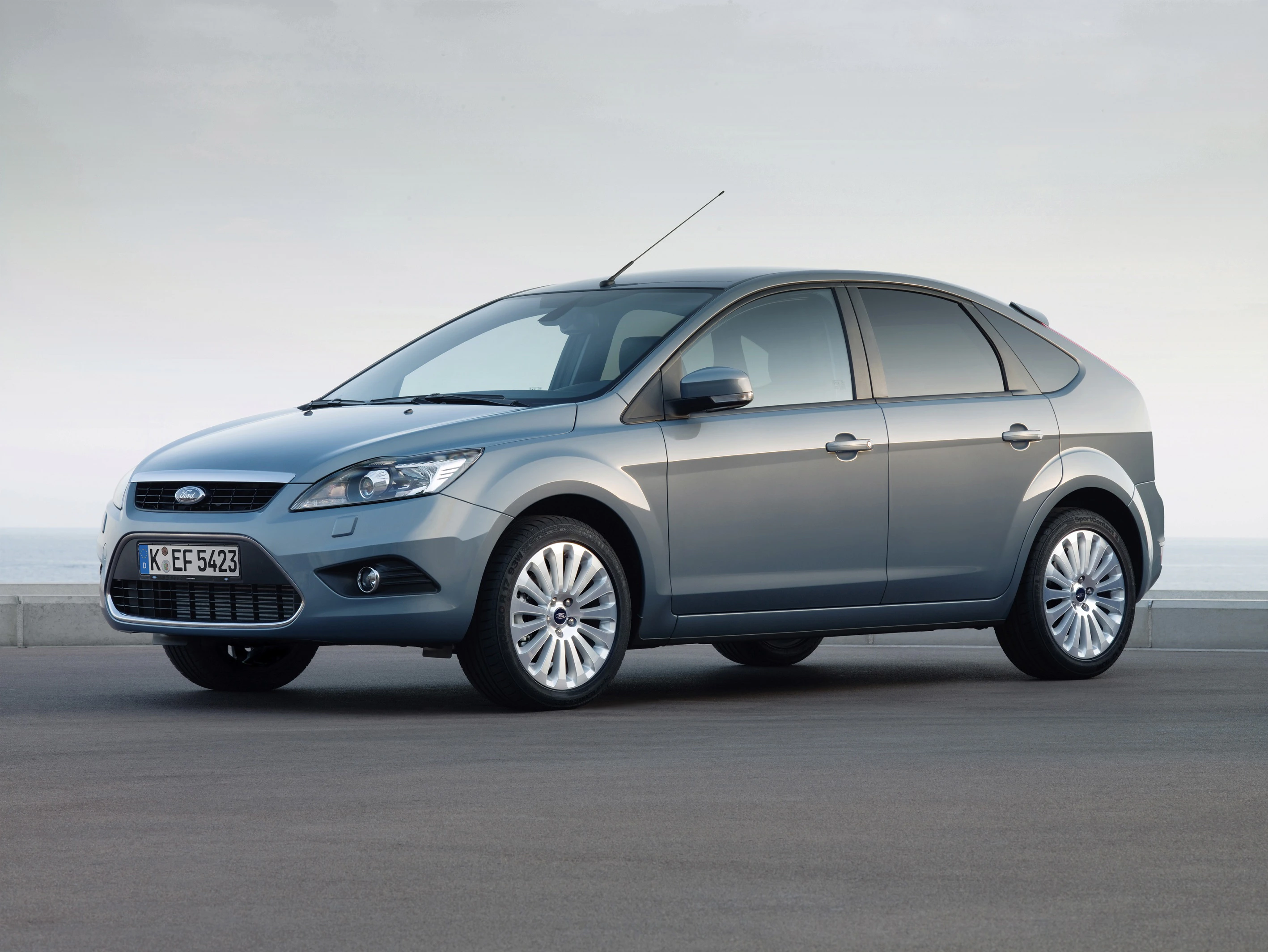 Grauer Ford Focus steht auf einer Straße vor einem bewölkten Himmel. Das Auto ist seitlich fotografiert, Reifen und Fenster sind sichtbar.