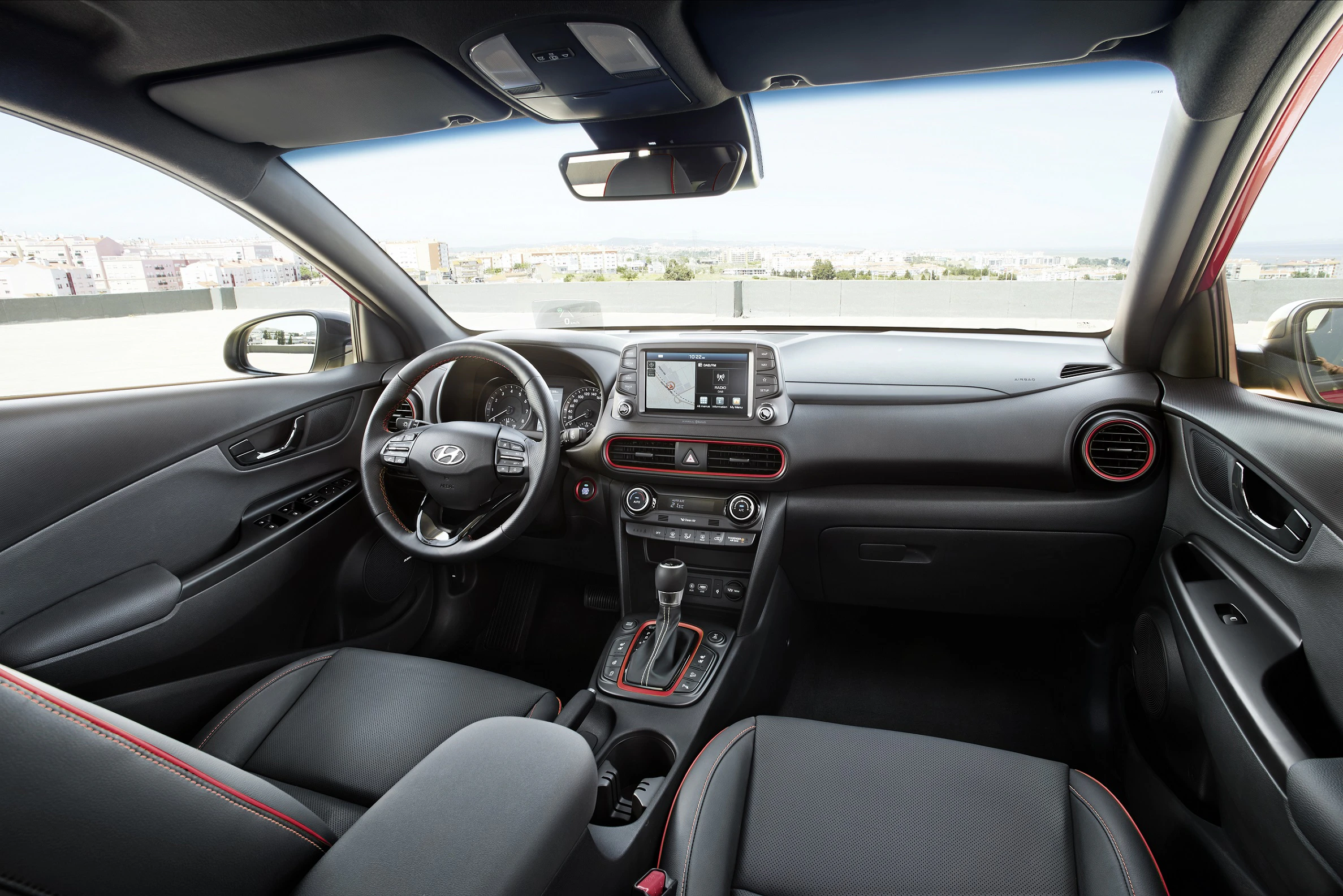 Moderne Innenansicht eines Hyundai-Kona-Autos, mit schwarze Ledersitzen, Lenkrad, Touchscreen-Display und Stadtblick durch die Windschutzscheibe.