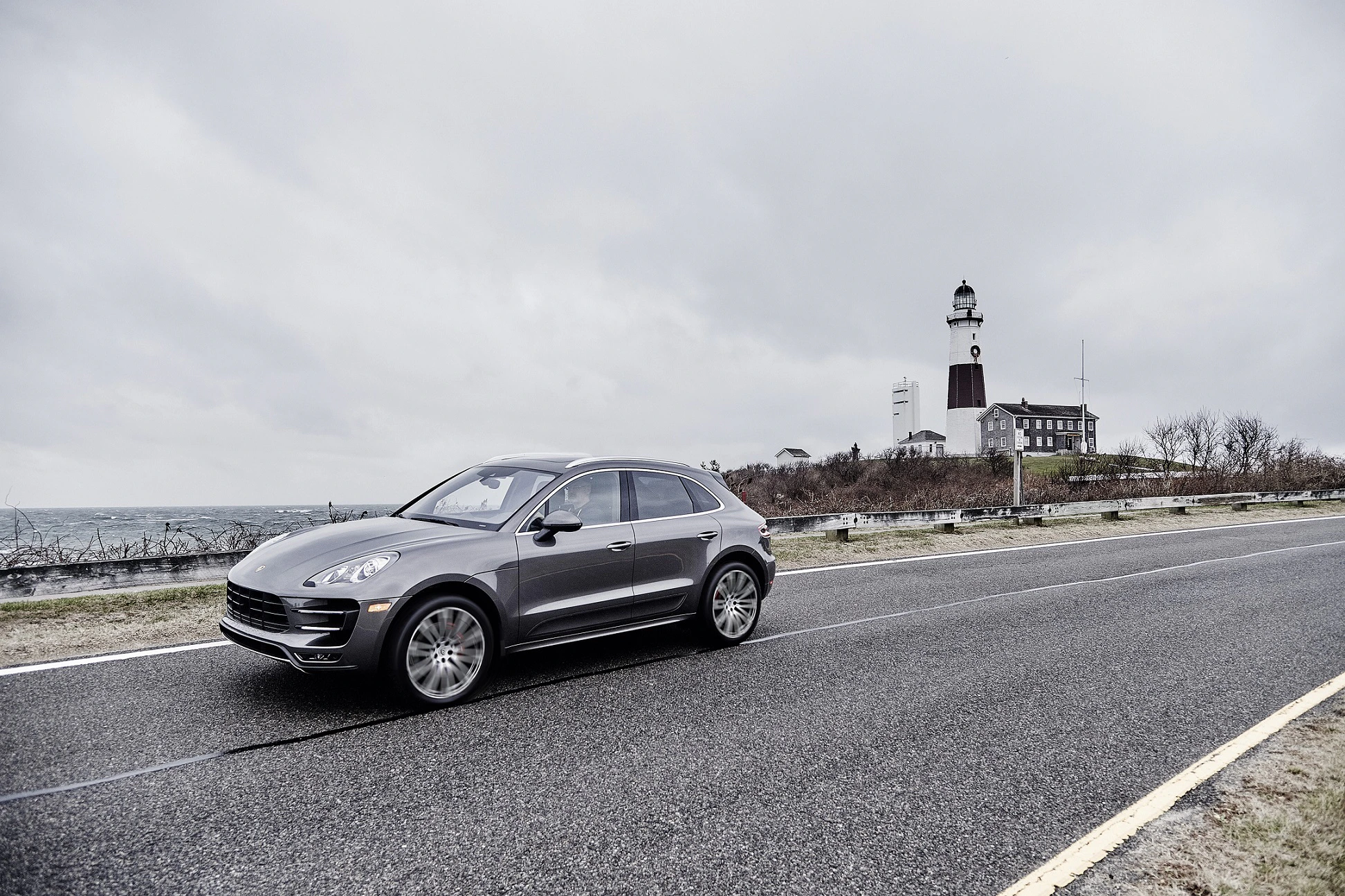 Grauer SUV fährt auf einer Küstenstraße, Leuchtturm im Hintergrund unter bewölktem Himmel.